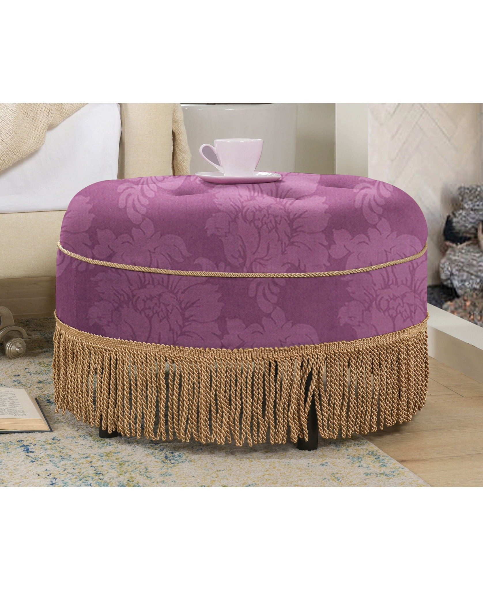 The Yolanda - Luxurious Upholstery Round Upholstered Accent Ottoman, featuring purple floral fabric and gold fringe trim, sits by a bed with an open book nearby, topped with a white teacup and saucer.