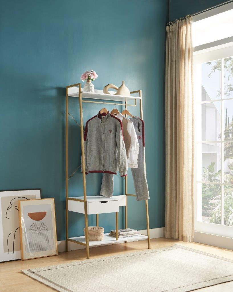 The Metal Clothes Rack Open Wardrobe Free Standing With One Drawer Hanging Clothes Rod displays two grey tracksuits, a vase of pink flowers, and a white pitcher. Nearby are framed art and books. Sunlight pours in through beige curtains.