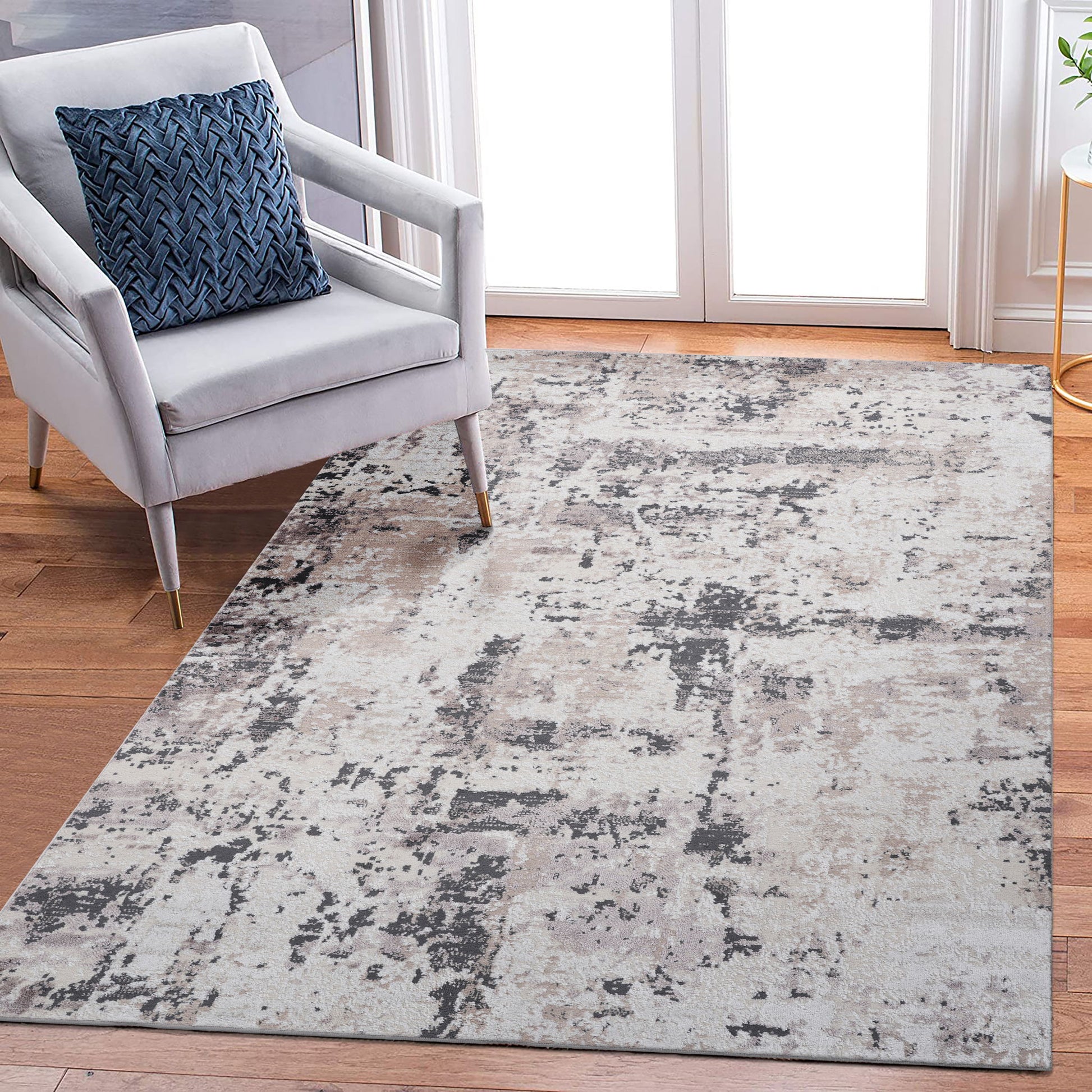 A modern living room with a light gray armchair and a blue textured cushion, featuring the Payas - Abstract Area Rug in gray tones on hardwood floors, and floor-to-ceiling windows in the background.