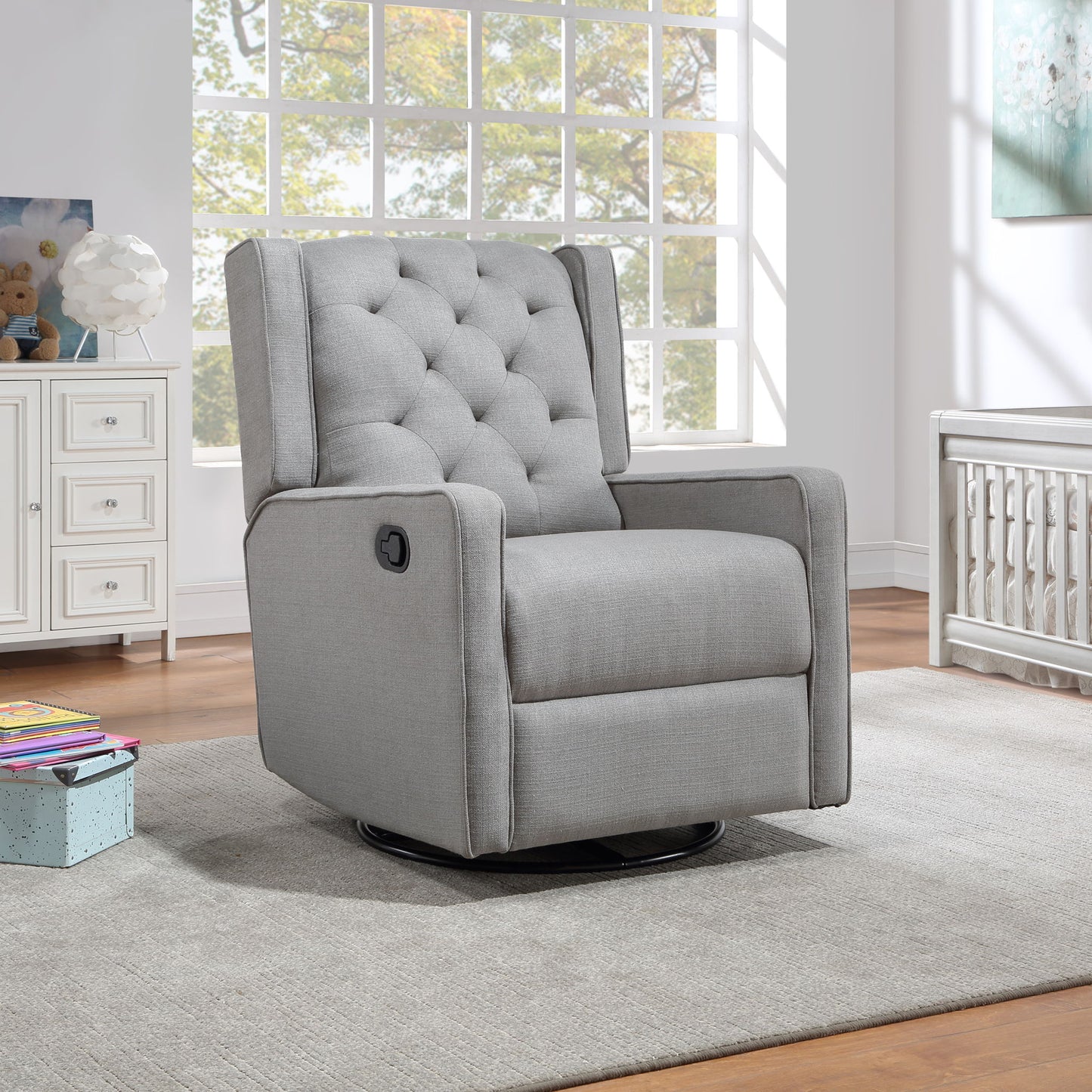 The Milah Gliding Swivel Recliner Tufted chair in light gray is on a gray rug in a bright room with large windows, next to a crib and white dresser. Books and a toy box are scattered nearby.