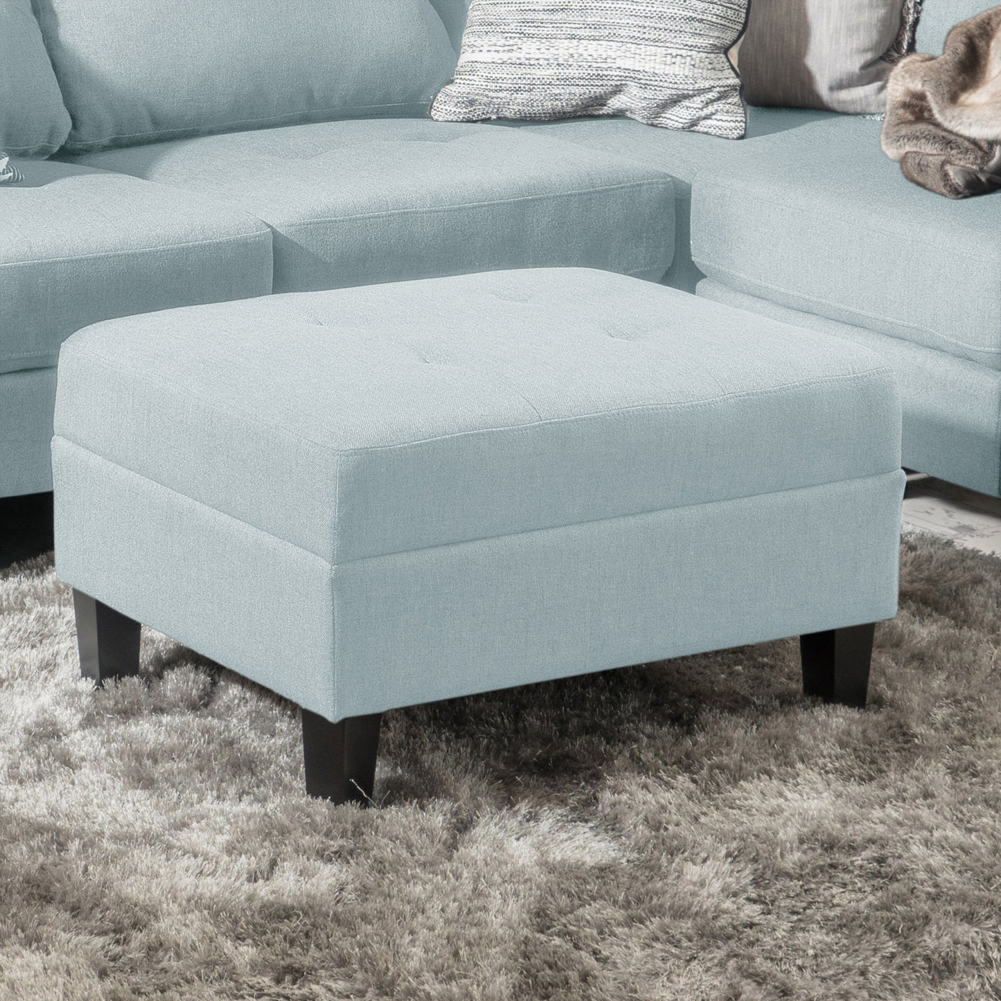 The Classic Ottoman With Birch Wood Legs And Fabric Upholstery, in light blue, sits on a gray shag rug before a matching sectional sofa with throw pillows.