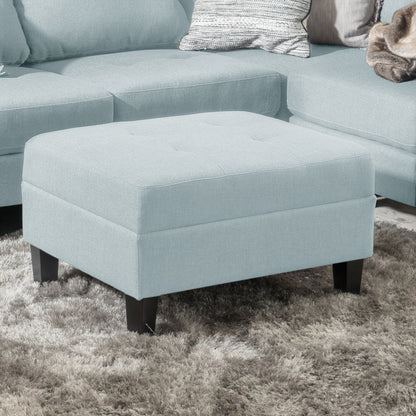 The Classic Ottoman With Birch Wood Legs And Fabric Upholstery, in light blue, sits on a gray shag rug before a matching sectional sofa with throw pillows.