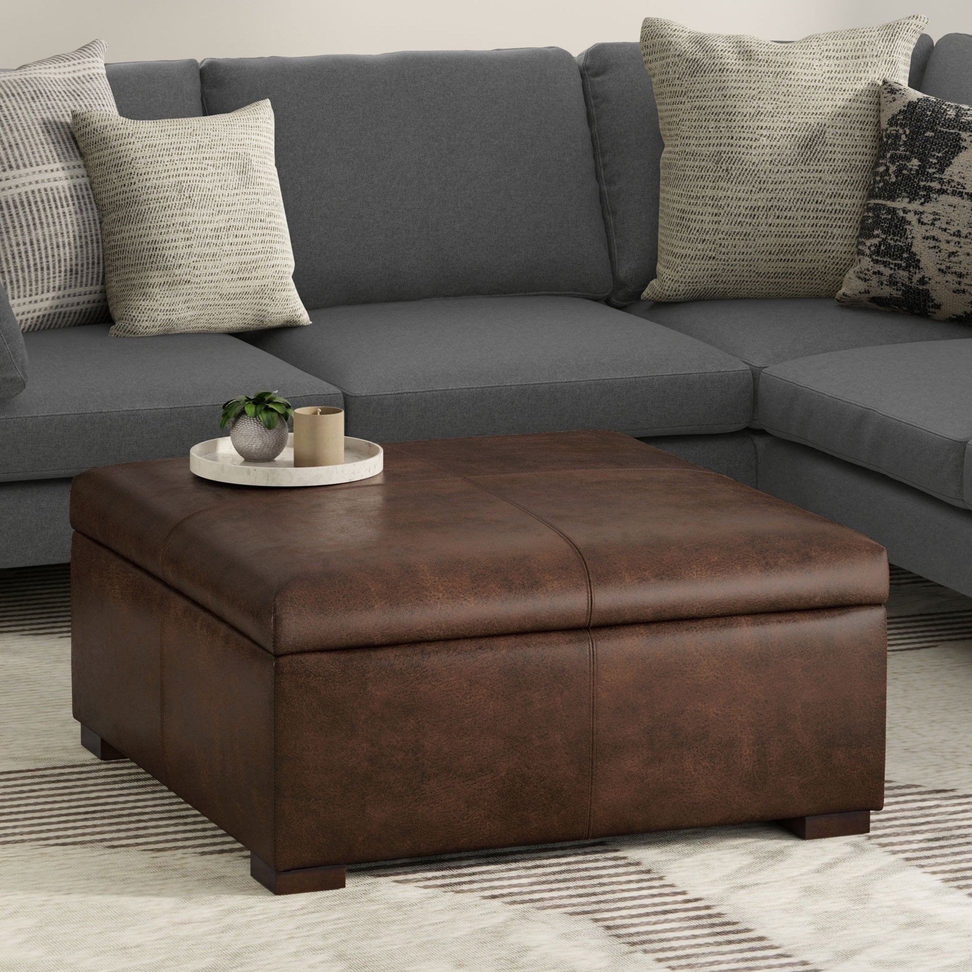 The Gabbie Coffee Table Upholstered Storage Ottoman sits on a cream and tan rug in front of a gray sectional with patterned pillows, topped with a round tray holding a small plant and books.