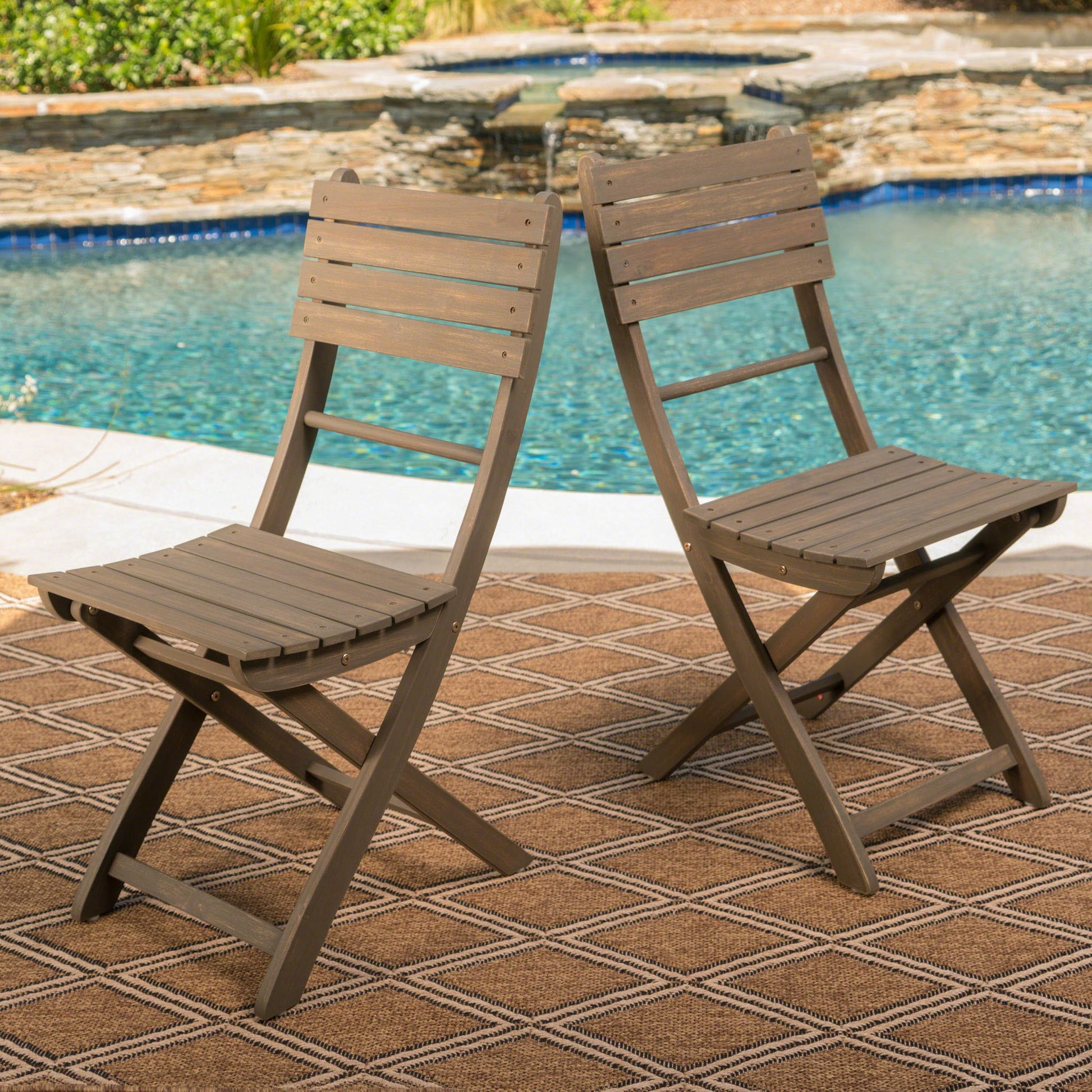 A set of two Outdoor Foldable Wood Dining Chairs with a slat design sit on a patterned outdoor rug beside a pool, surrounded by stone landscaping and lush greenery.