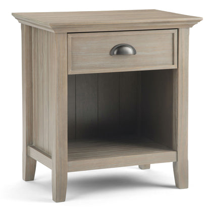 The Acadian Handcrafted Bedside Table features light wood, a single drawer with a metal handle, an open lower shelf, simple modern design, slightly tapered legs, and vertical grooves on the side panels.