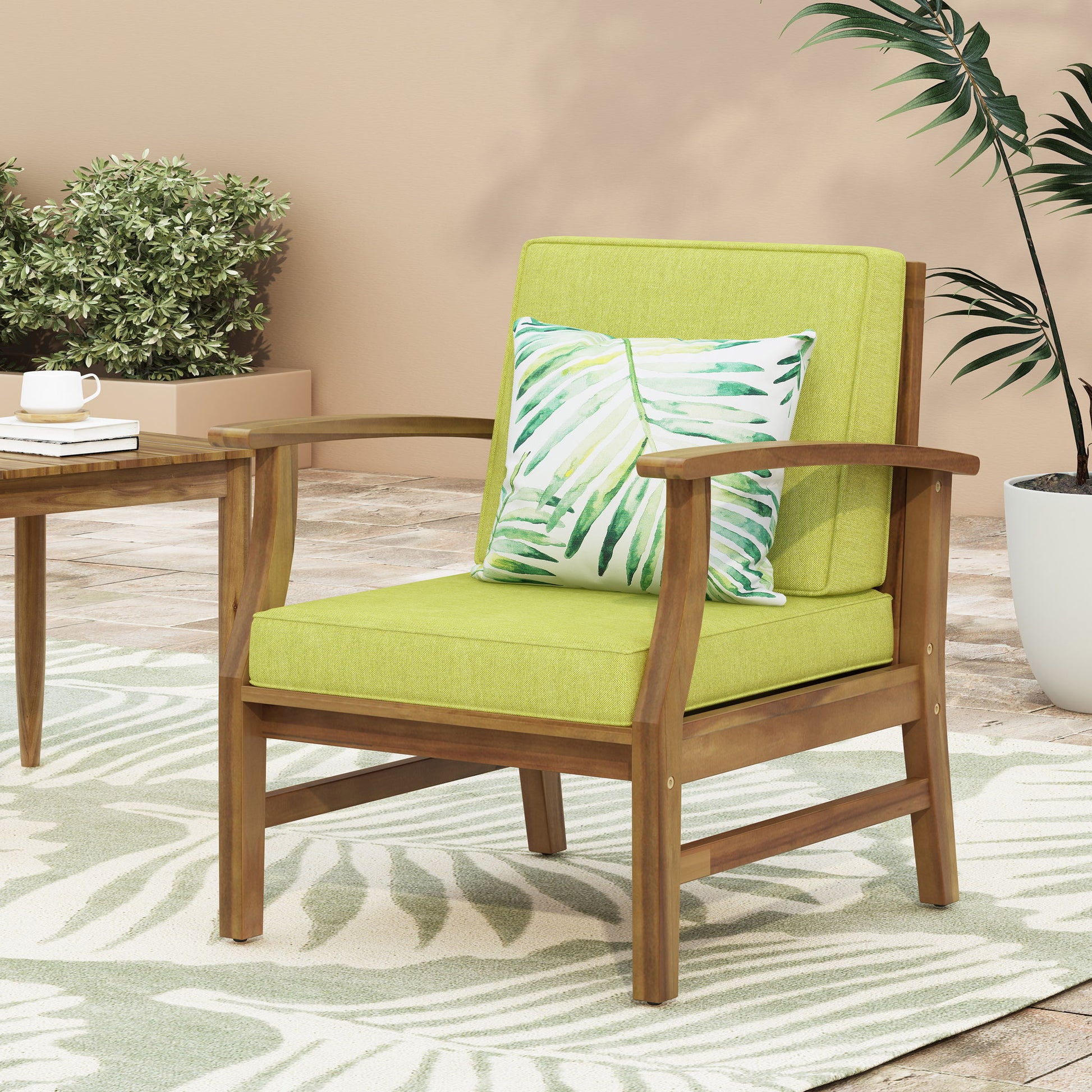 The Perla Contemporary Club Chair with water-resistant cushions, paired with a matching table, sits on the patio among leafy plants and a light-colored leaf-patterned rug.