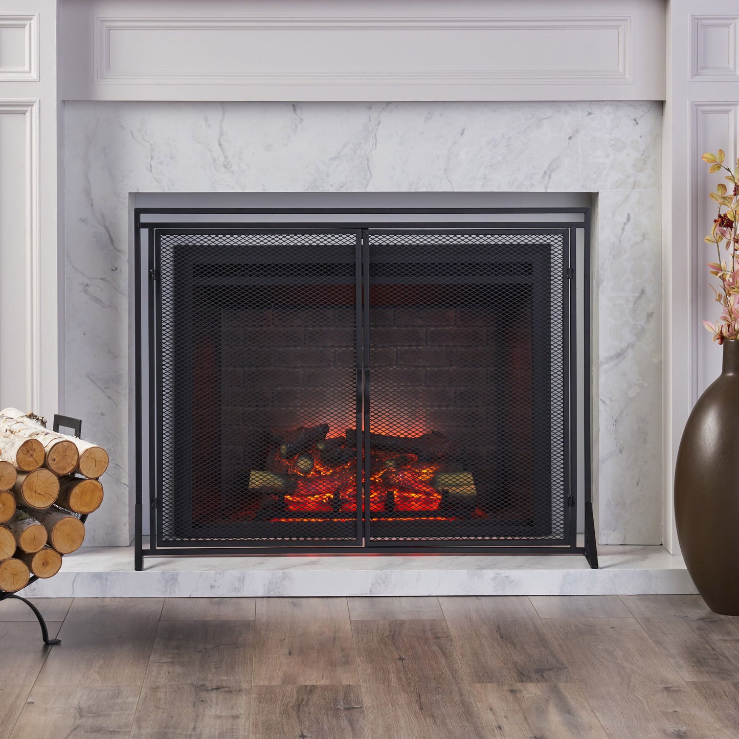 A modern fireplace features the Samantha Iron Fireplace Screen with a 2-door design, glowing logs and embers, a white marble surround, firewood stacked in a log holder to the left, and a tall brown vase with flowers to the right on a wooden floor.