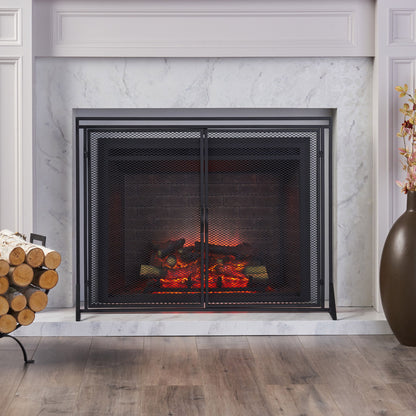 A modern fireplace features the Samantha Iron Fireplace Screen with a 2-door design, glowing logs and embers, a white marble surround, firewood stacked in a log holder to the left, and a tall brown vase with flowers to the right on a wooden floor.