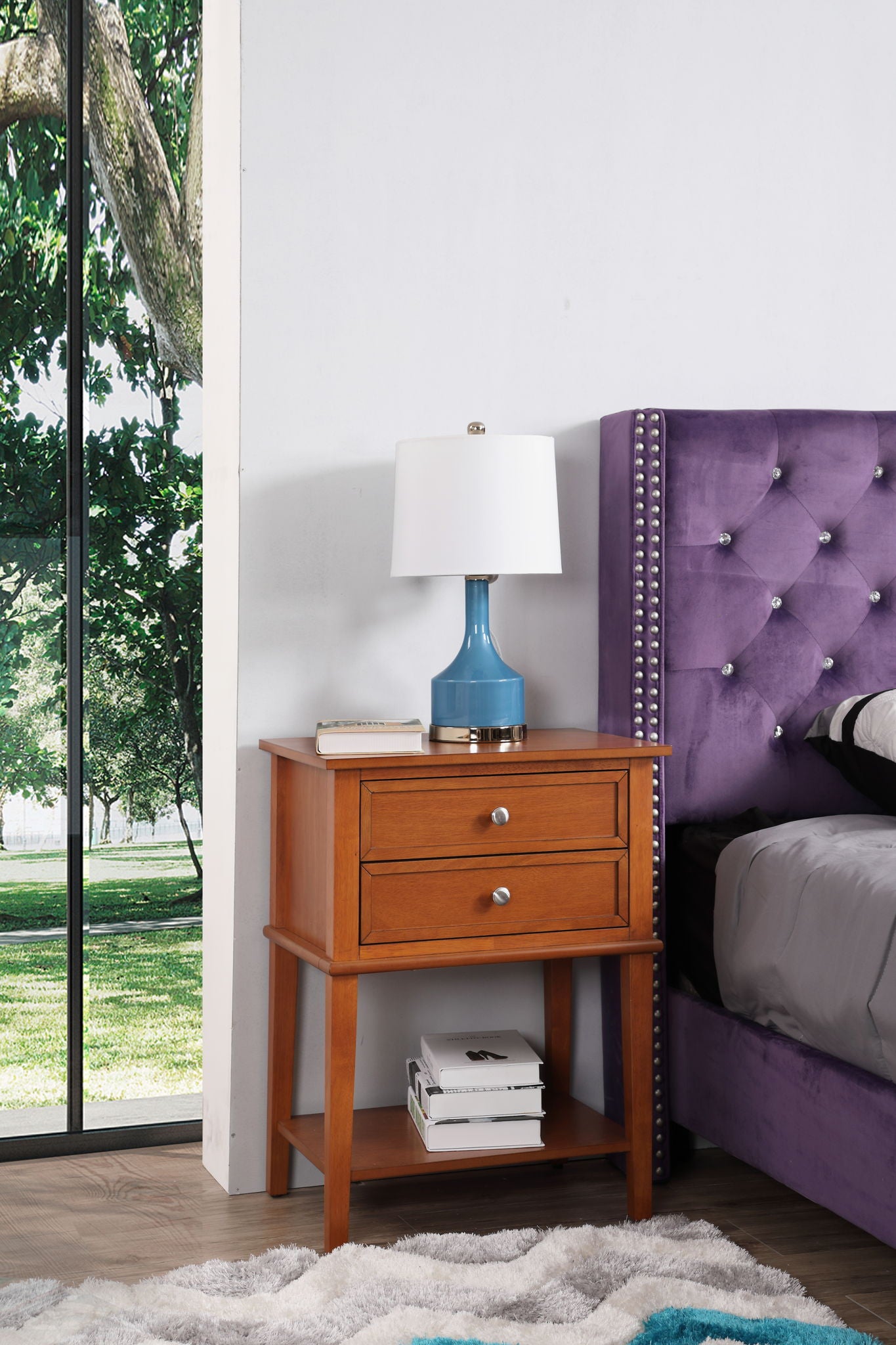 The Vibrant Contemporary Nightstand with two drawers and a lower shelf for books stands beside a purple upholstered bed. A blue lamp rests on the nightstand, while a large window reveals lush trees and greenery outside.