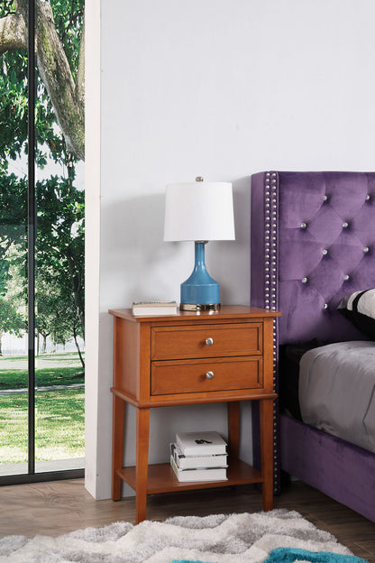 The Vibrant Contemporary Nightstand with two drawers and a lower shelf for books stands beside a purple upholstered bed. A blue lamp rests on the nightstand, while a large window reveals lush trees and greenery outside.