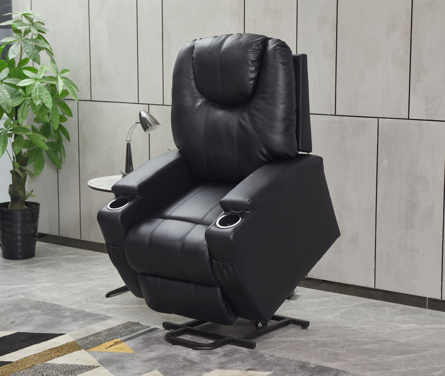 The Modern Recliner Chair With Cup Holders and remote control in black leather features a raised footrest, shown in a stylish room with a potted plant, small side table, and wall lamp nearby.