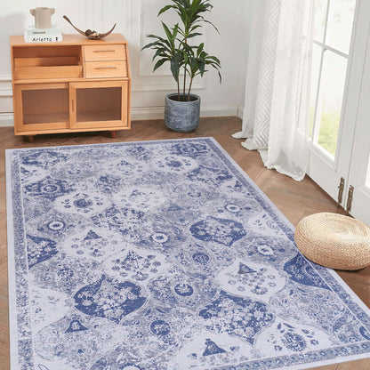 The Maval - Stunning Area Rug, featuring a blue and white pattern, is displayed on a wooden floor beside a woven pouf, potted plant, wooden cabinet, and glass doors draped with white curtains that let in natural light.
