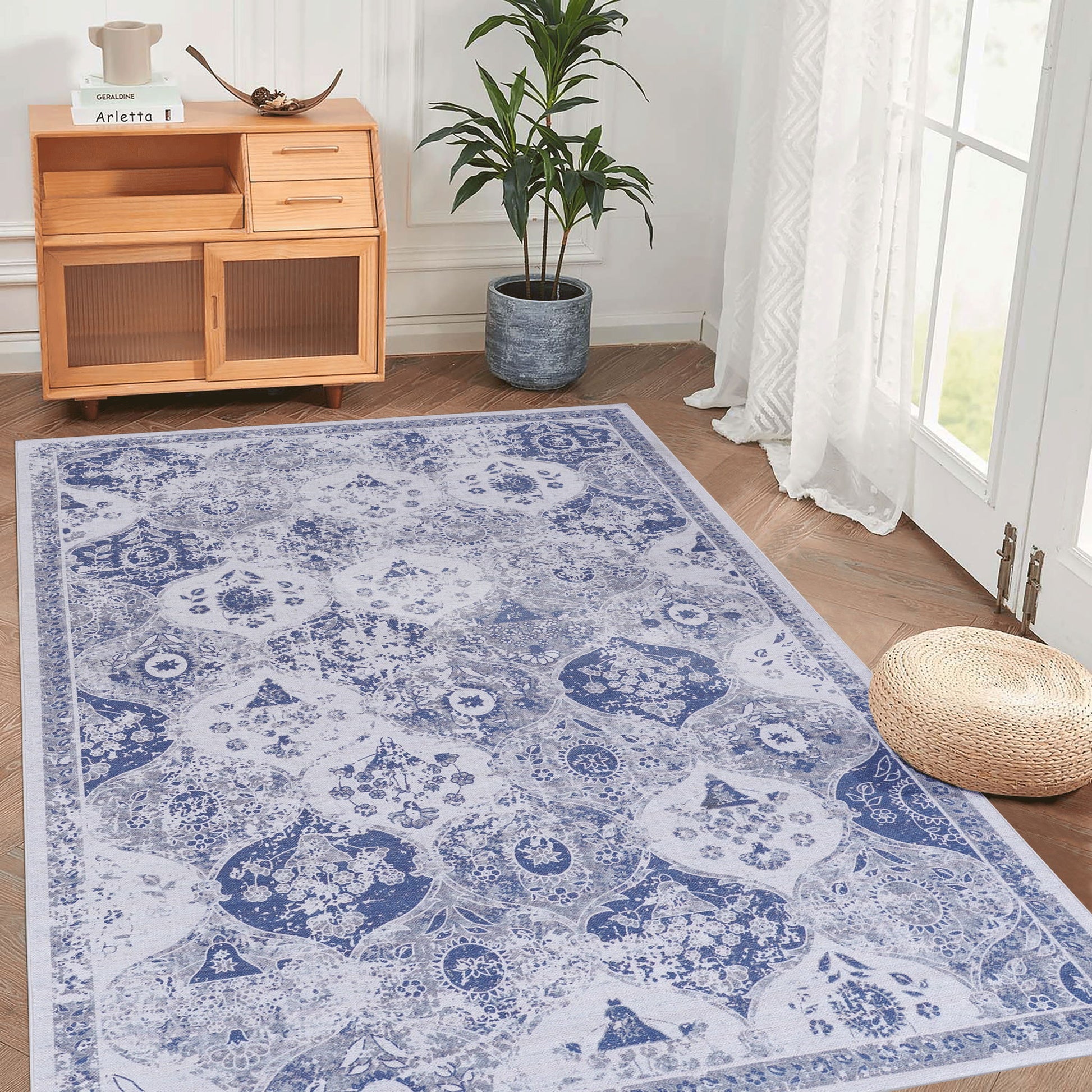 A room featuring the Maval - Area Rug in blue and white on a wooden floor, a small wooden cabinet with shelves, a potted plant, a round woven pouf, and glass doors with white curtains letting in natural light.