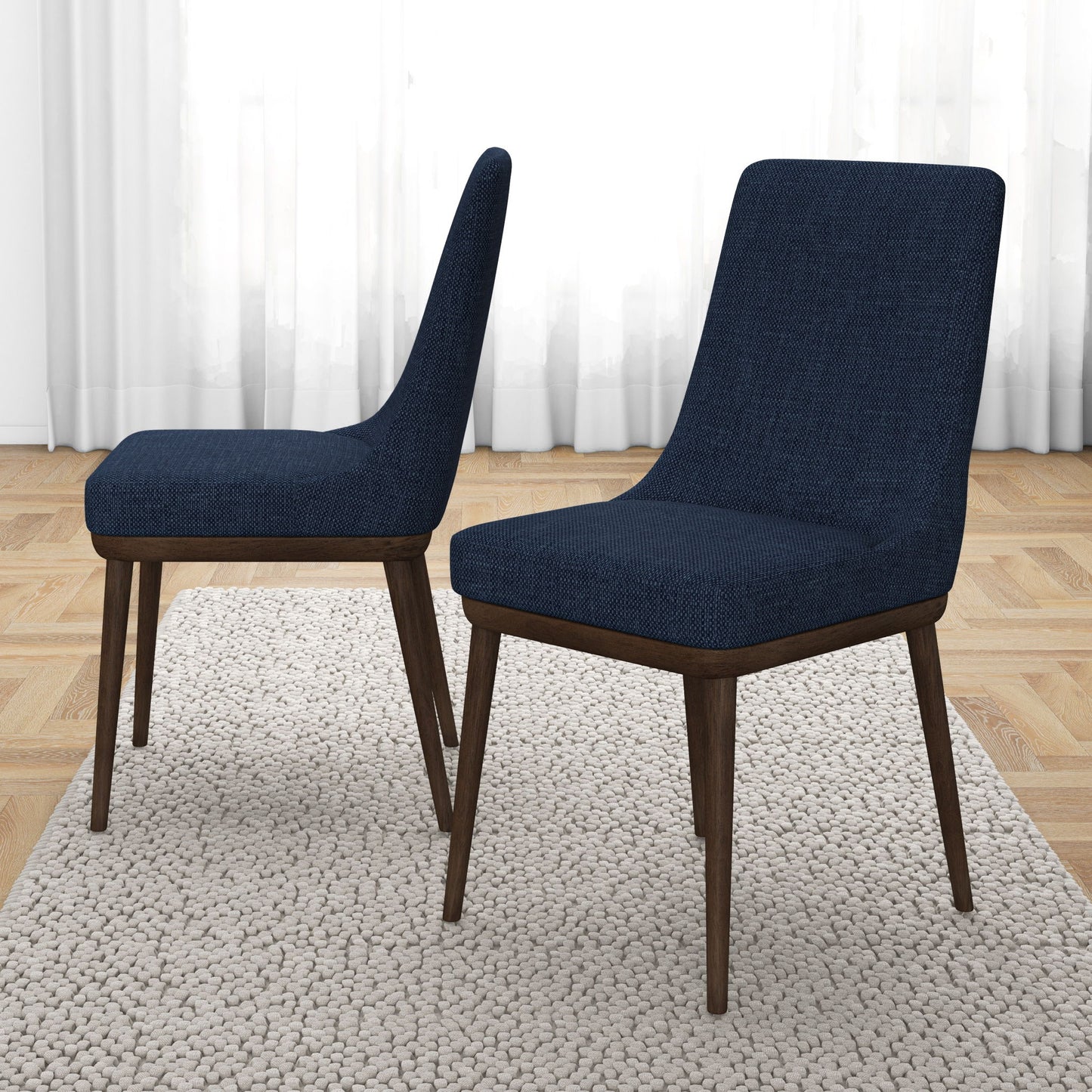 The Kate Mid-Century Modern Dining Chairs (Set of 2), featuring navy blue upholstery and dark wood legs, are set on a textured light rug in a room with parquet flooring and white curtains.