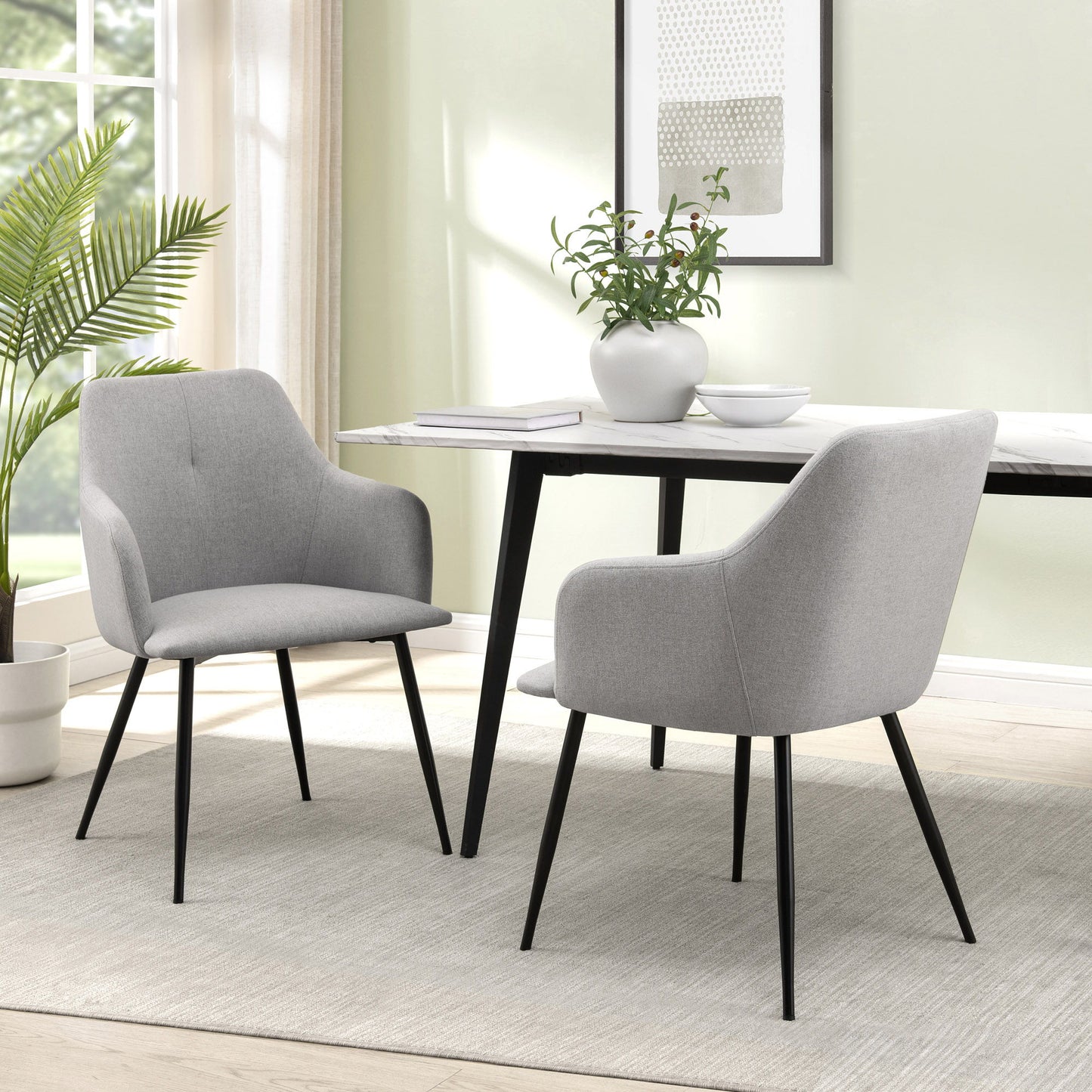 Two Contemporary Upholstered Woven Dining Chairs with black legs are placed at a rectangular white table in a bright room featuring a large window, potted plant, wall art, and a neutral rug.