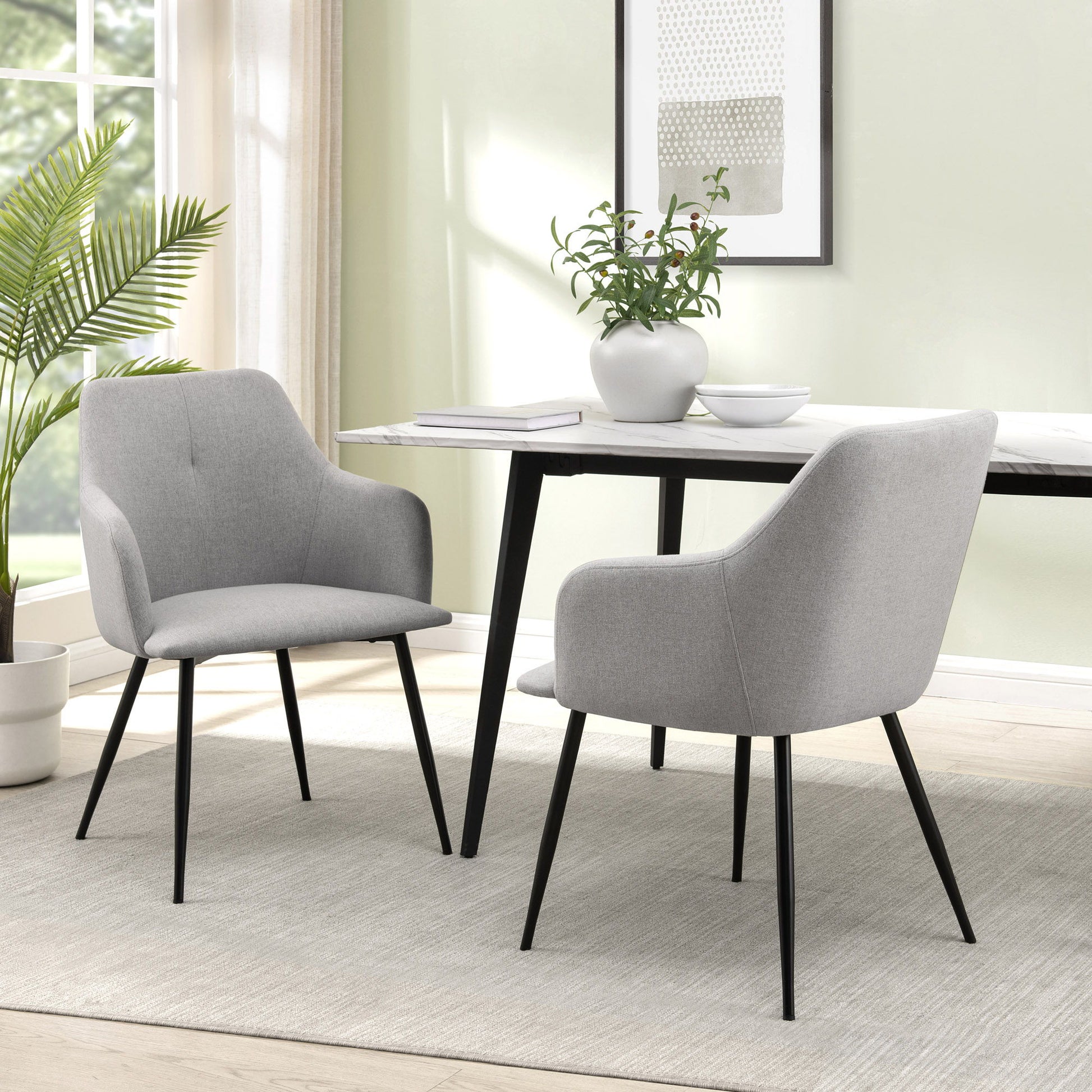 Two Contemporary Upholstered Woven Dining Chairs with black legs are placed at a rectangular white table in a bright room featuring a large window, potted plant, wall art, and a neutral rug.