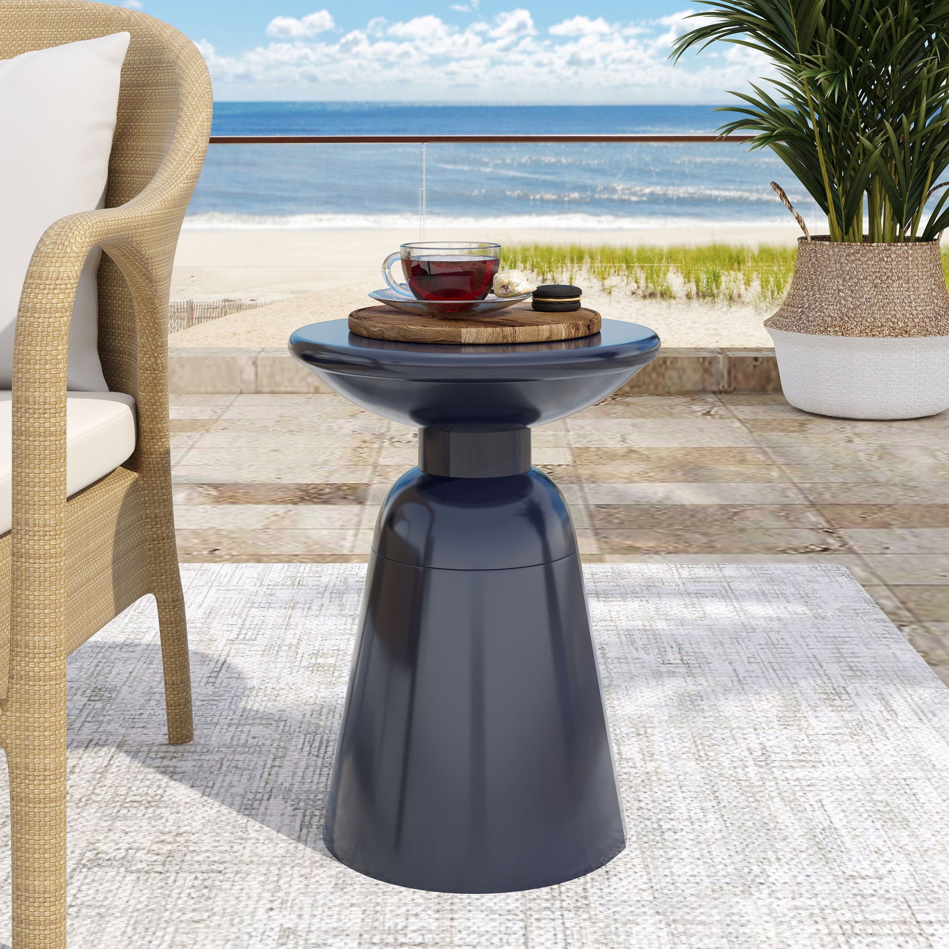 On a patio, the Outdoor Modern Metal End Large Table holds a wooden tray with a glass teacup and saucer. In the background, there’s a wicker chair, green plant, and a sandy beach with ocean waves.