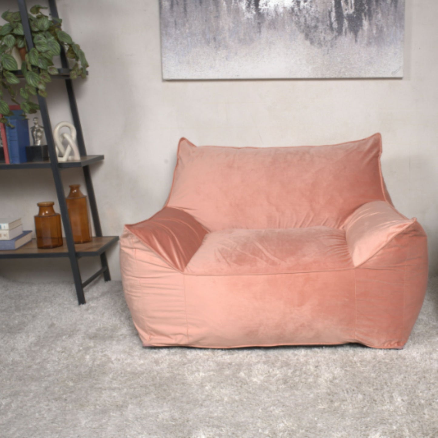 The Allea - Bean Bag Chair With Armrests, in large pink plush, sits on a light gray rug in a modern living room with a bookshelf, potted plant, and abstract wall art in the background.