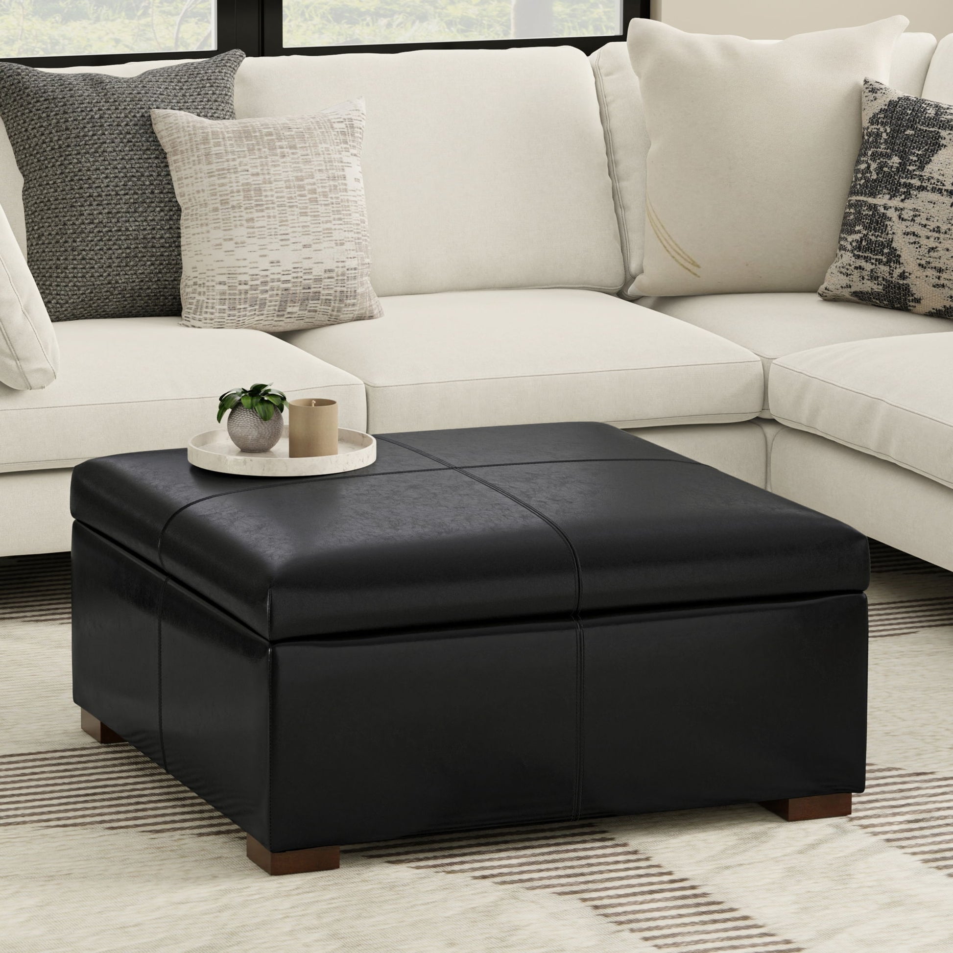 The Gabbie Coffee Table Upholstered Storage Ottoman in black stands before a cream sectional sofa with patterned pillows. A round tray holding a plant and candle tops the ottoman, complementing the room’s modern neutral decor.