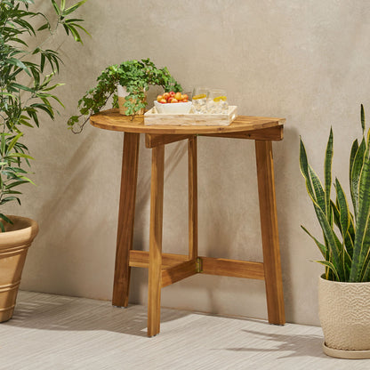 An Outdoor Half Round Folding Acacia Wood Bistro Table holds a tray with two glasses and a bowl of cherry tomatoes, surrounded by potted green plants against a beige wall and light floor.