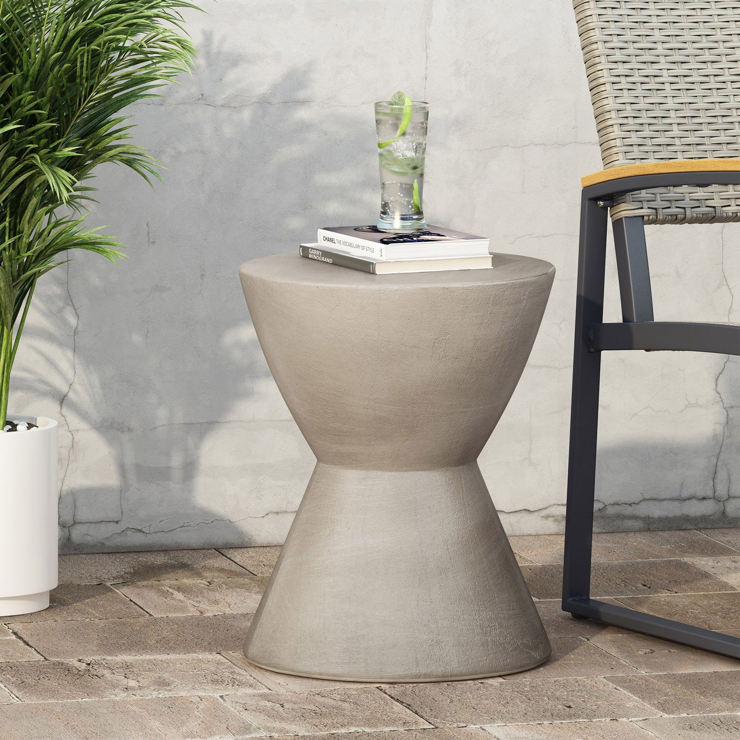 The Outdoor Hourglass Shaped Side Table in light gray sits on a patio beside a wicker chair and potted plant, holding a glass of lime water and a book against a textured wall.