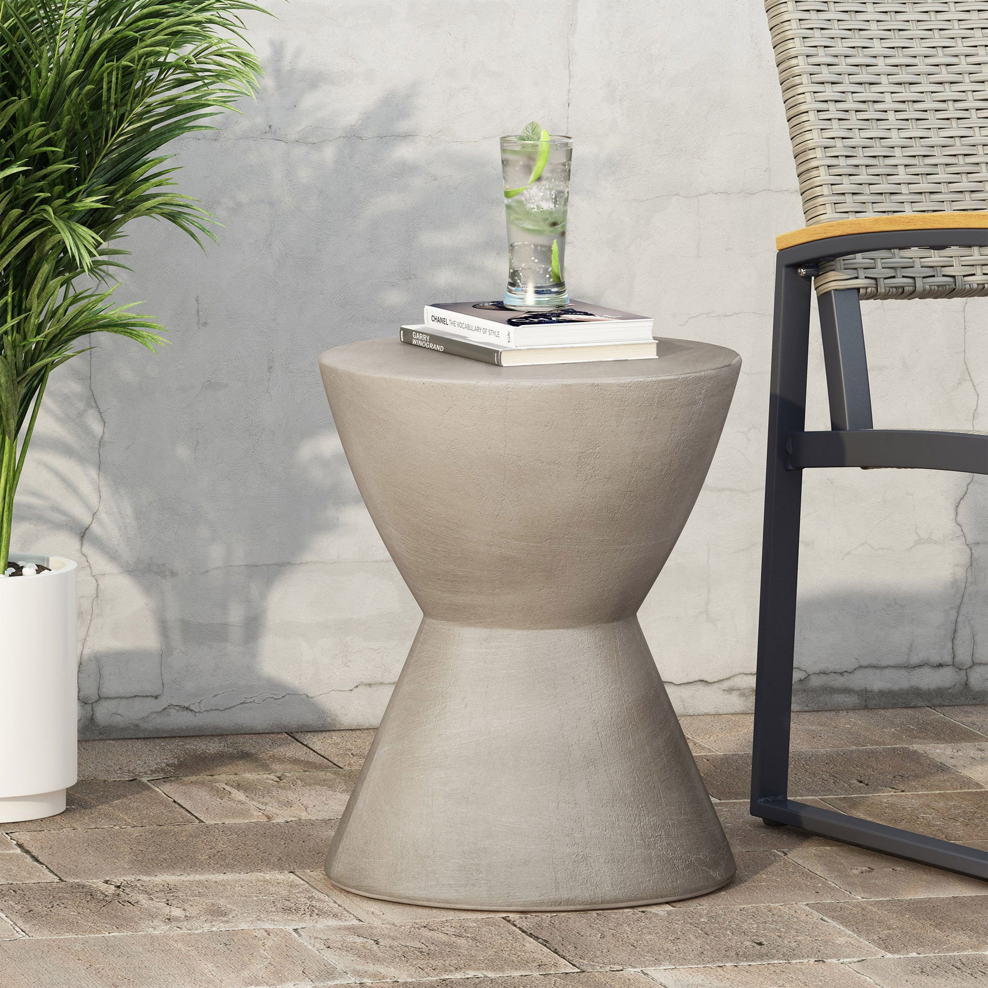 The Outdoor Hourglass Shaped Side Table in light gray sits on a patio beside a wicker chair and potted plant, holding a glass of lime water and a book against a textured wall.