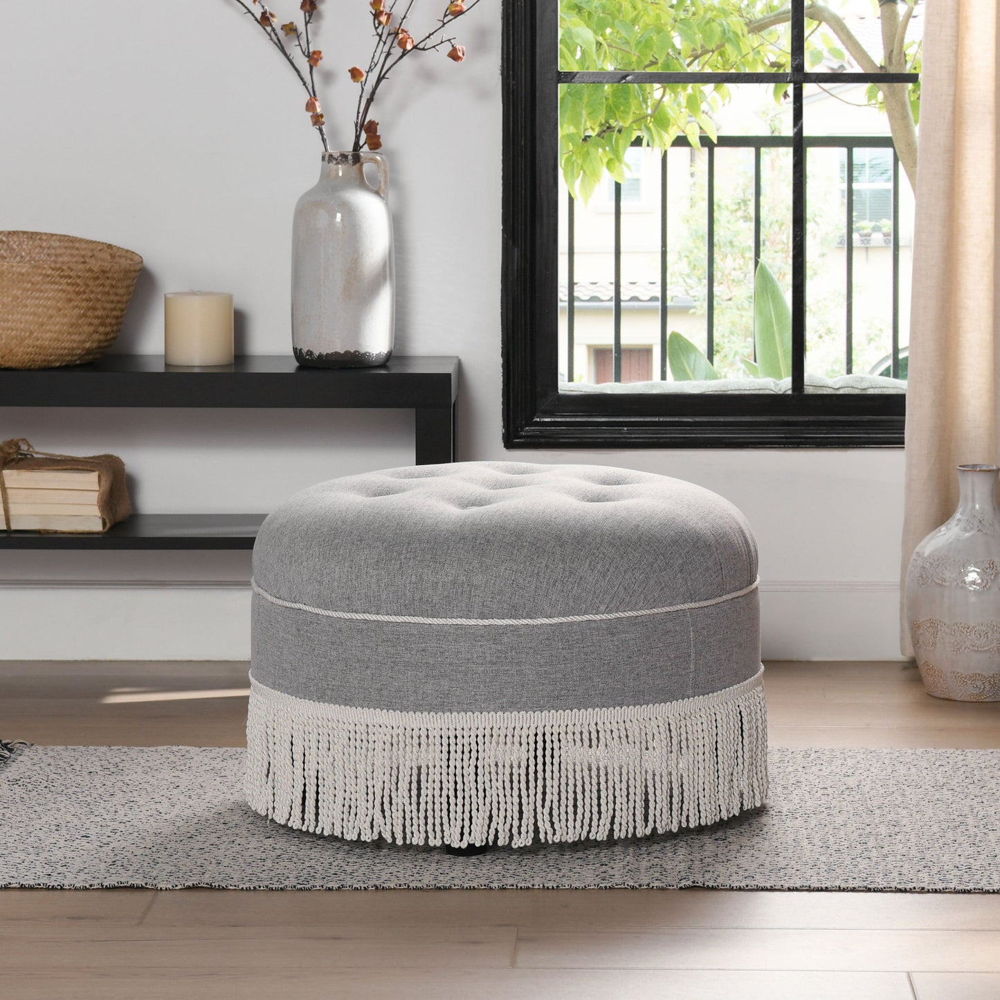 The Yolanda - Round Upholstered Accent Ottoman in gray with white fringe sits on a rug in a bright living room near a window, with vases, a basket, and a candle displayed on a nearby shelf and floor.