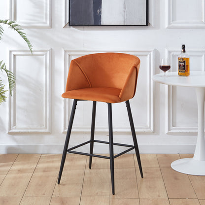 A set of two modern orange Counter Height Bar Stools with curved backs and black metal legs stands on a wooden floor beside a white table holding a bottle, glass, and framed artwork against a classic white paneled wall.