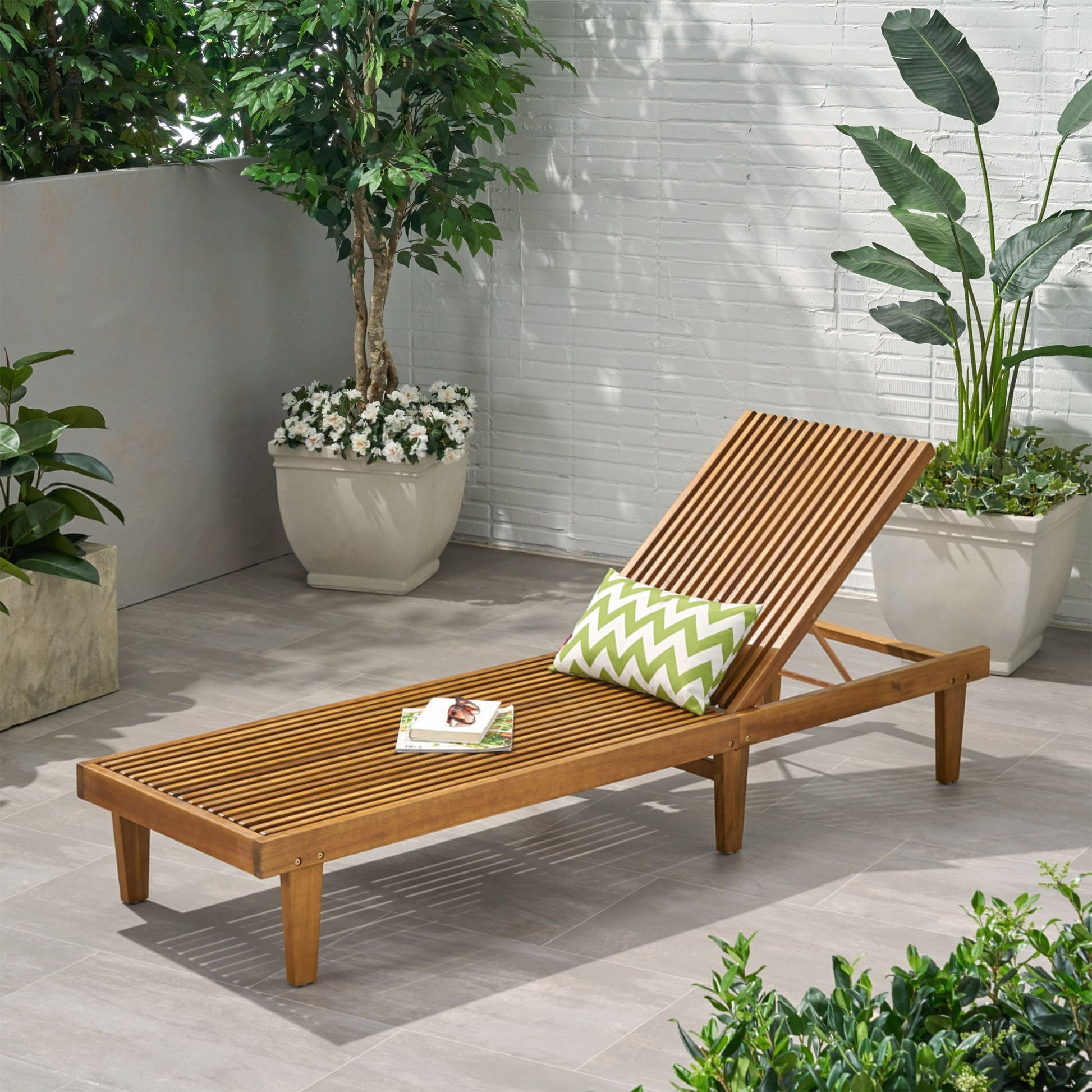 The Nadine - Stylish Chaise Lounge Chair - Teak, topped with a green and white chevron pillow and a book, rests on a tiled patio amid lush potted plants against a white brick wall.