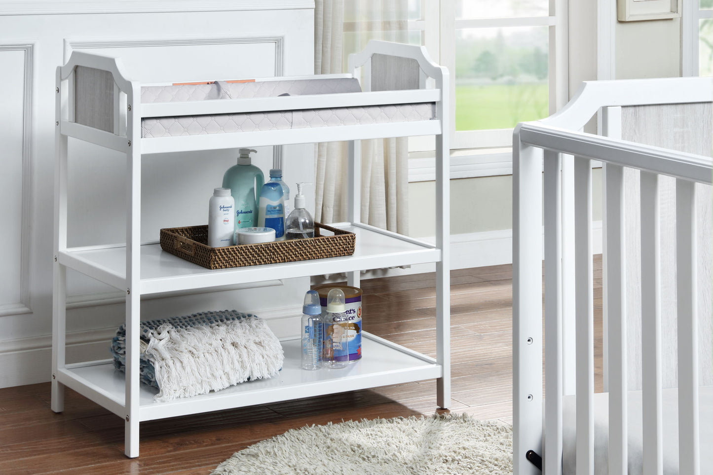 The Brees - Changing Table, finished in white, features shelves stocked with bottles, lotion, a basket, and folded blankets. It is placed next to a white crib in a bright, softly lit nursery.