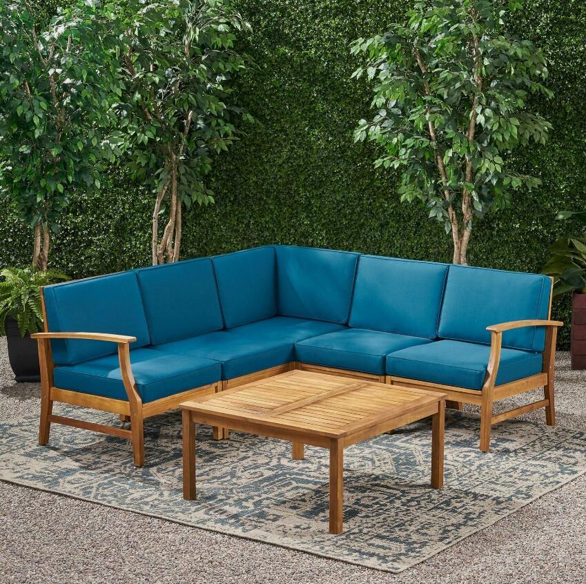 The Perla - 6 Pieces Sofa Set features an L-shaped wooden outdoor sofa with blue cushions surrounding a square coffee table, all arranged on a patterned rug with green hedges and trees in the background.
