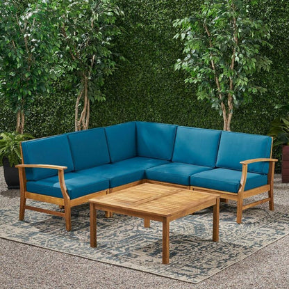 The Perla - 6 Pieces Sofa Set features an L-shaped wooden outdoor sofa with blue cushions surrounding a square coffee table, all arranged on a patterned rug with green hedges and trees in the background.