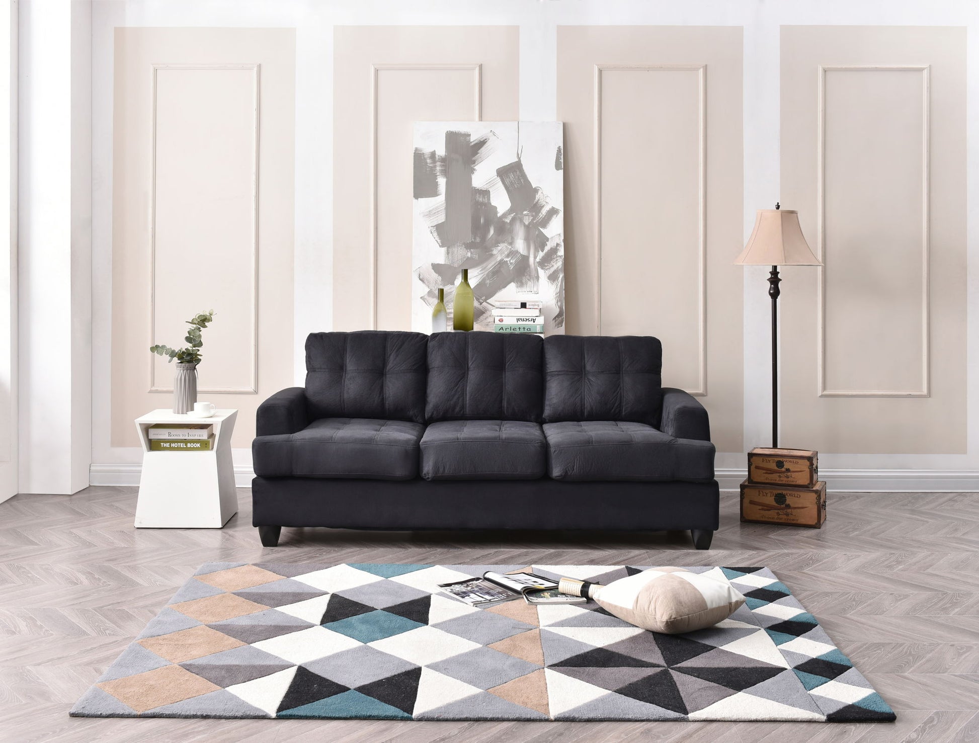 A modern living room features a dark gray Sandridge Sofa, a white side table with a plant and books, a geometric rug, large abstract painting, floor lamp, stacked vintage suitcases, and a pillow on the rug.