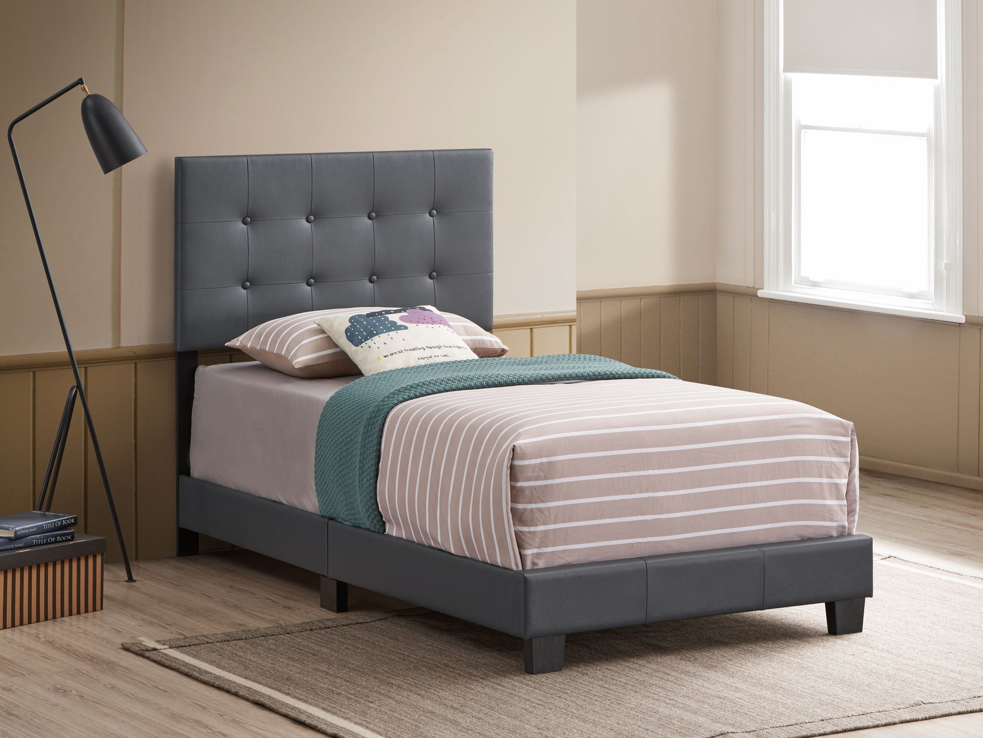 The Caldwell - Bed, featuring a gray tufted headboard and striped bedding with a teal throw, is set on a beige rug in a bright, minimalist bedroom accented with wood, a black floor lamp, and a large window.