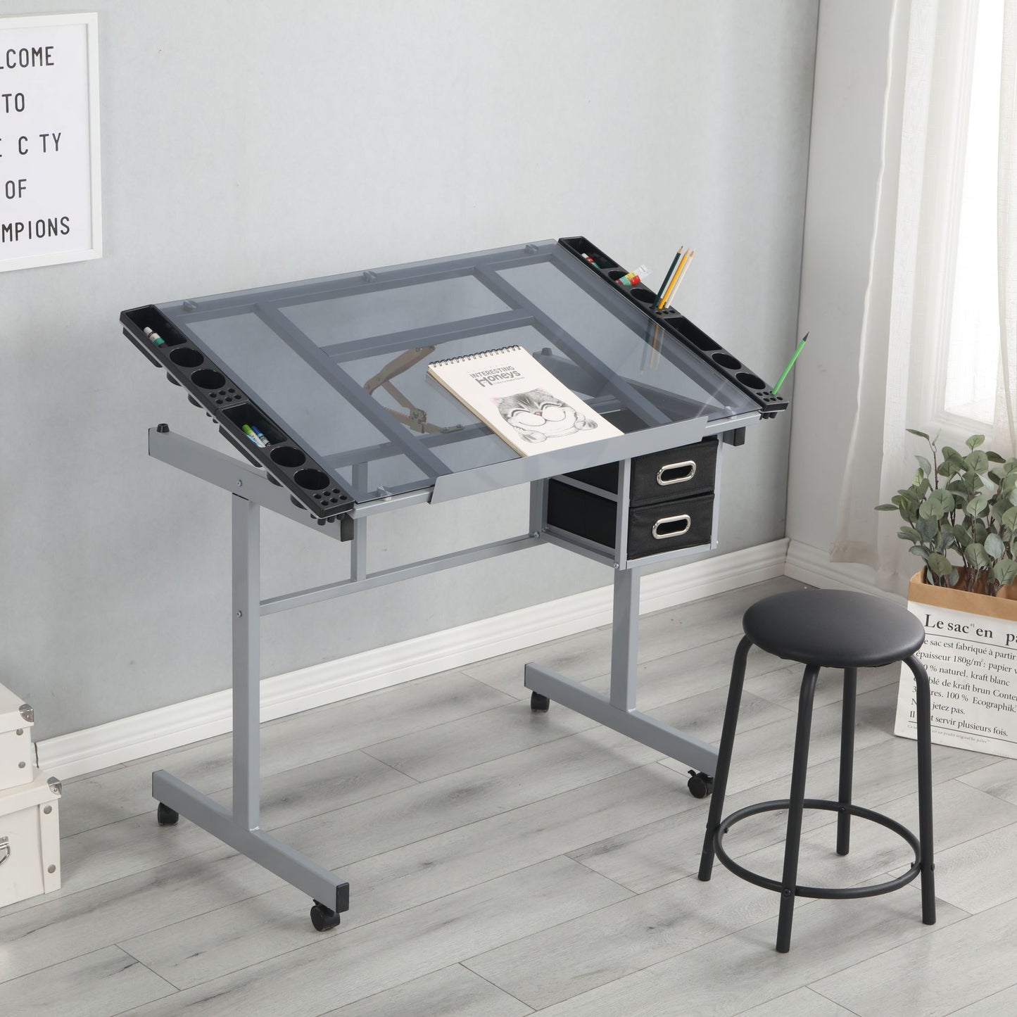 The Adjustable Art Drawing Desk Craft Station Drafting With 2 Non-Woven Fabric Slide Drawers And 4 Wheels - Silver is paired with a black stool on a light wood floor by a window with plants, featuring an angled glass top and art supplies.