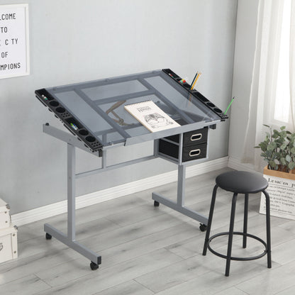 The Adjustable Art Drawing Desk Craft Station Drafting With 2 Non-Woven Fabric Slide Drawers And 4 Wheels - Silver is paired with a black stool on a light wood floor by a window with plants, featuring an angled glass top and art supplies.