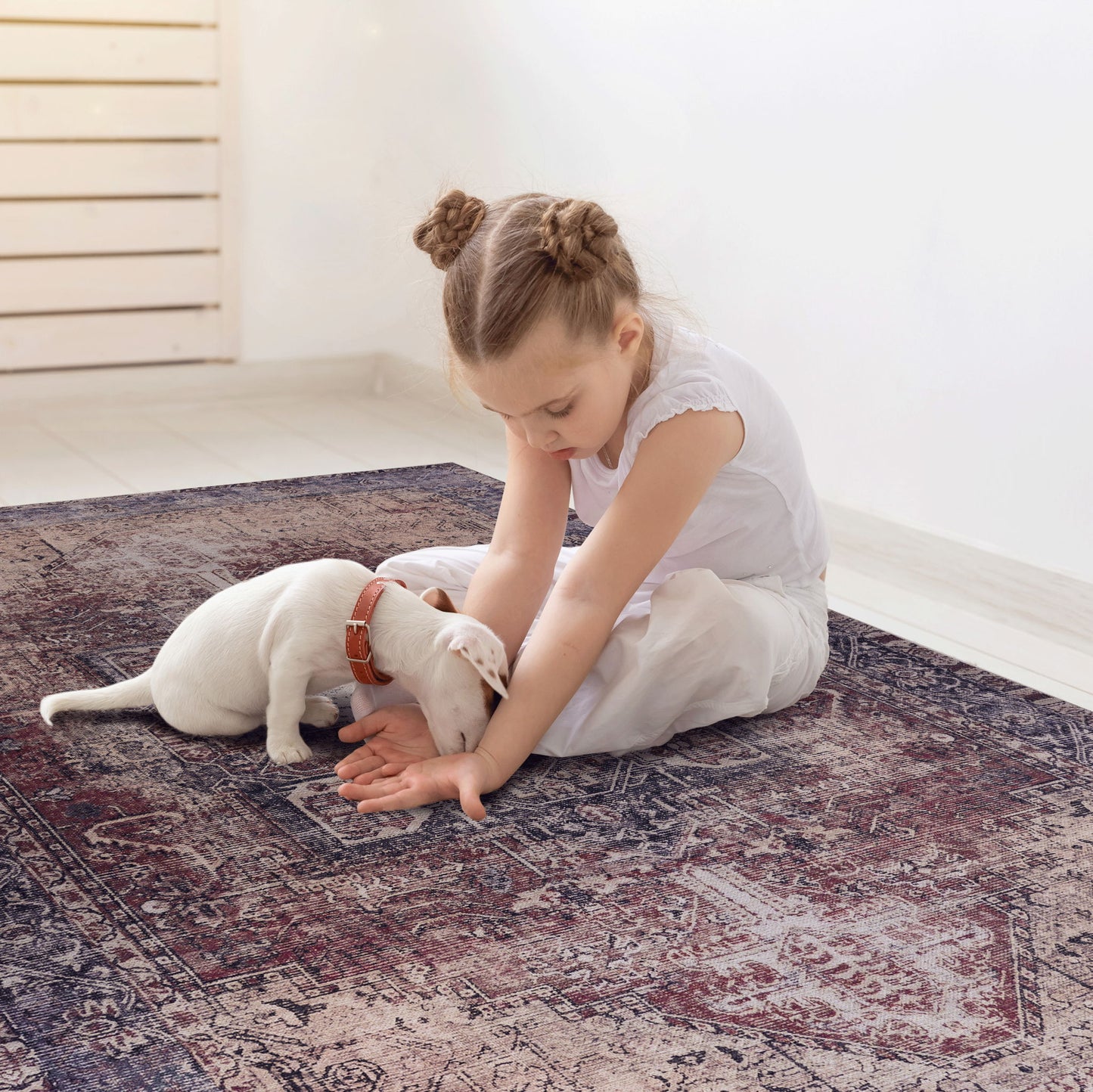 A young girl in a white dress sits on the Maval - Unique Washable Area Rug, gently petting a small white puppy with a brown collar. The cozy indoor scene features soft lighting and a light-colored wall in the background.