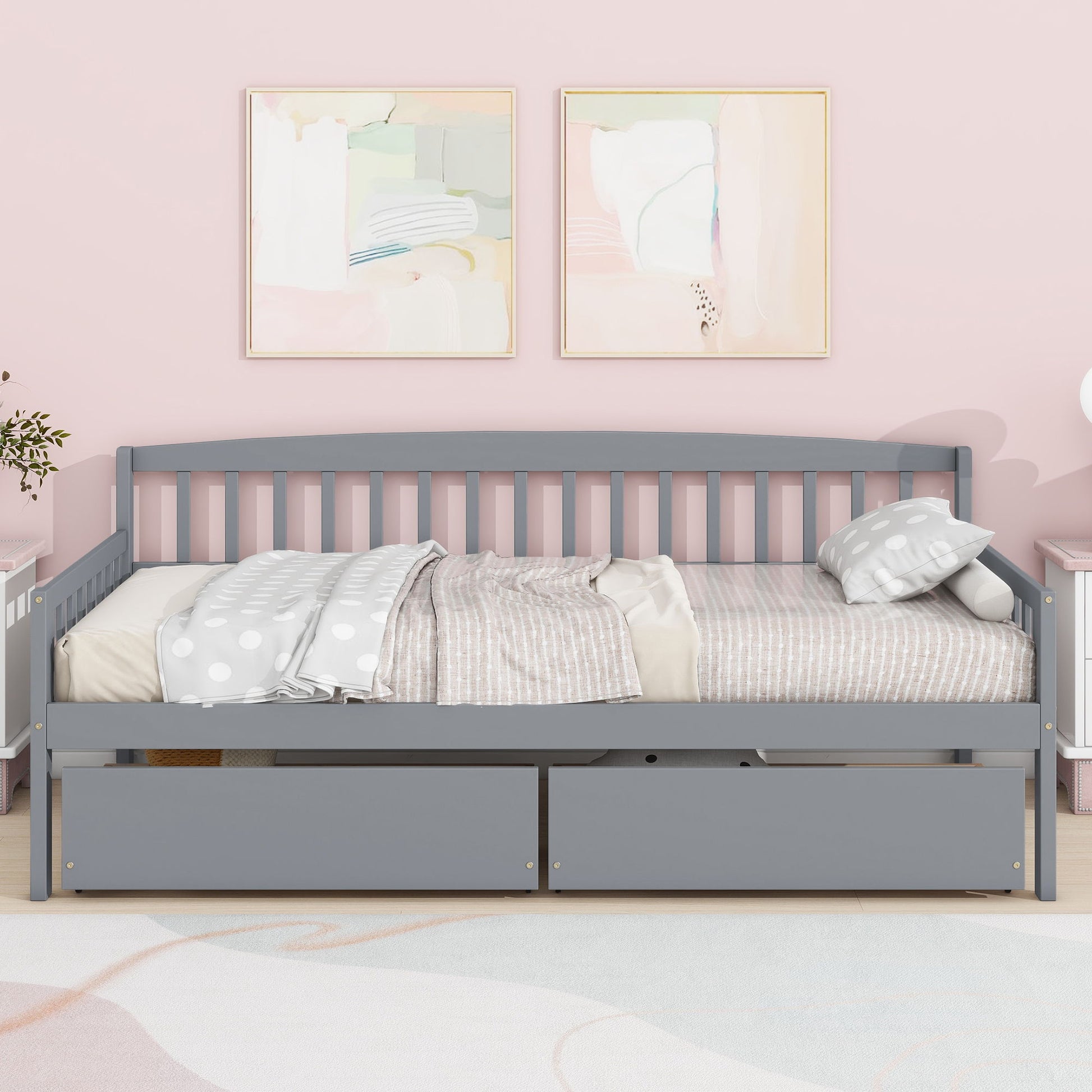 A Pine Wood Daybed with Two Storage Drawers, featuring a bed platform of 10 support slats, sits against a pink wall under two pastel abstract paintings. It’s dressed with a striped comforter and polka-dot blanket; white furniture and a light rug are nearby.