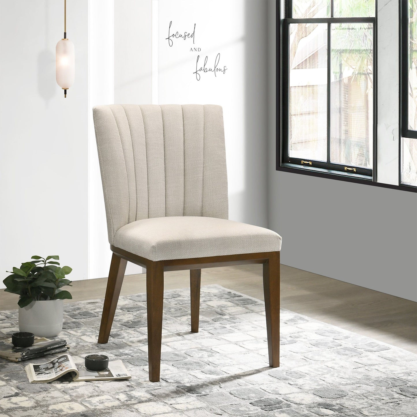 Two Elmwood Stylish Dining Chairs with beige upholstery and wooden legs sit on a patterned rug near a potted plant and window, sunlight streaming in. A wall sign nearby reads "focused and fabulous.