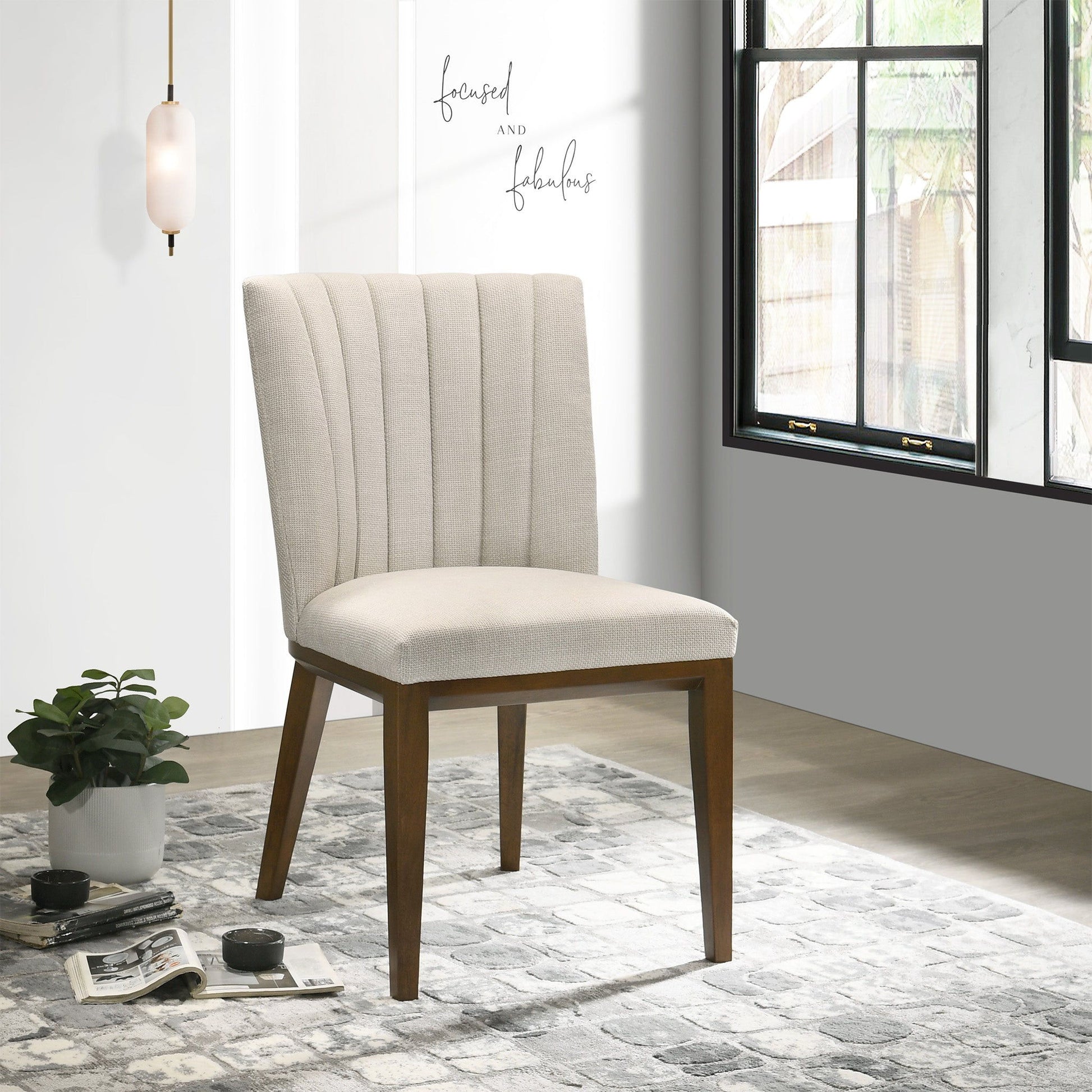 Two Elmwood Stylish Dining Chairs with beige upholstery and wooden legs sit on a patterned rug near a potted plant and window, sunlight streaming in. A wall sign nearby reads "focused and fabulous.