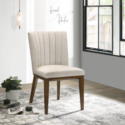 Two Elmwood Stylish Dining Chairs with beige upholstery and wooden legs sit on a patterned rug near a potted plant and window, sunlight streaming in. A wall sign nearby reads "focused and fabulous.