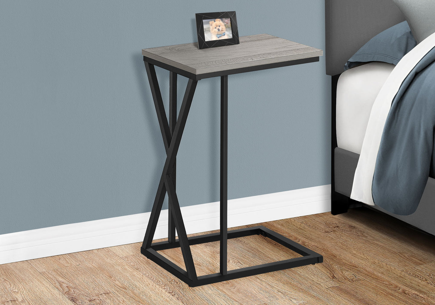 The Accent Table C - Shaped, Contemporary & Modern with a gray wood top and black metal frame sits by a bed, topped with a framed dog photo. The room features wood flooring and blue-gray walls.
