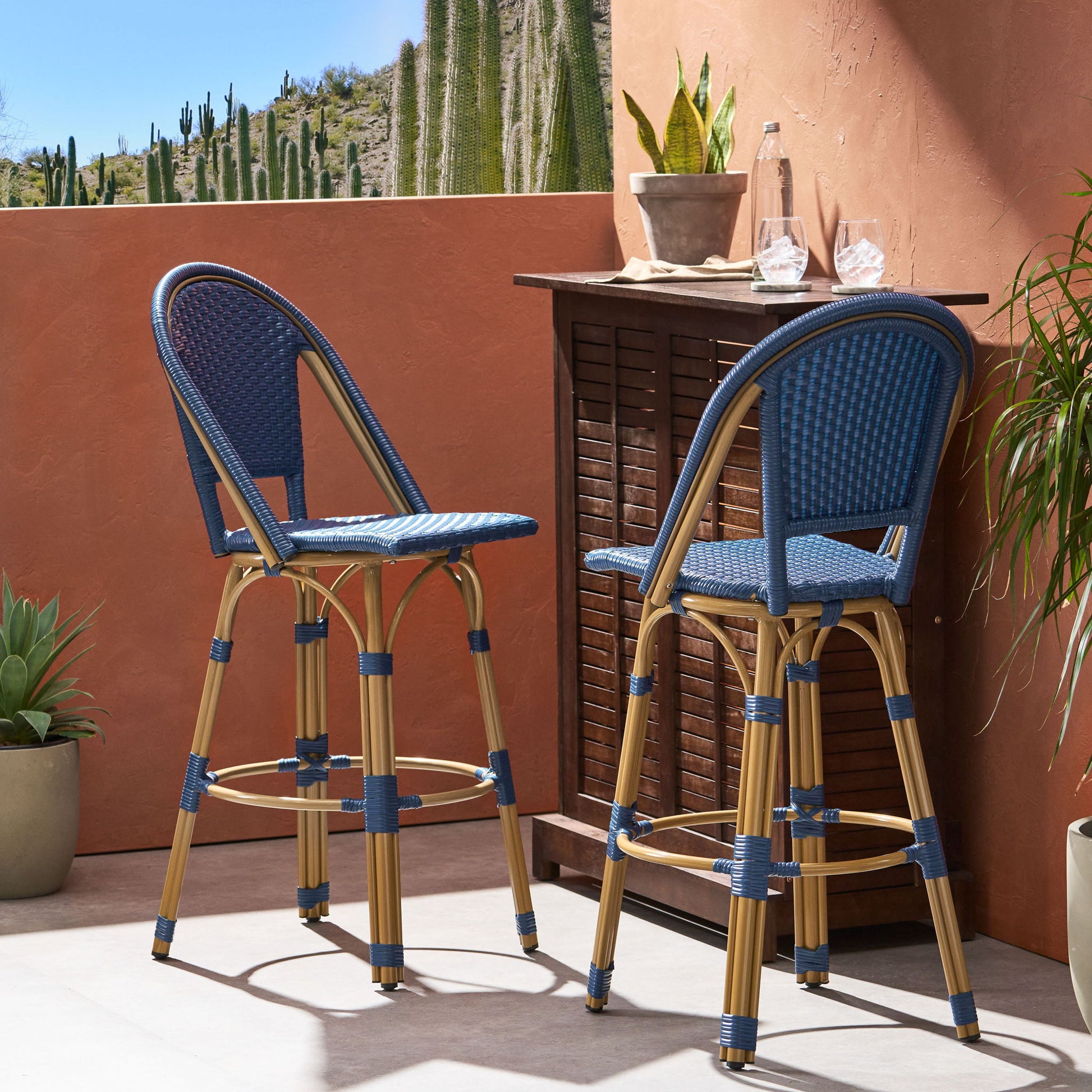 Two Outdoor Rattan And Aluminum French Barstools sit by a wooden bar table on a sunny patio with potted plants and cacti. Two glasses, a clear carafe, and a small plant decorate the table.