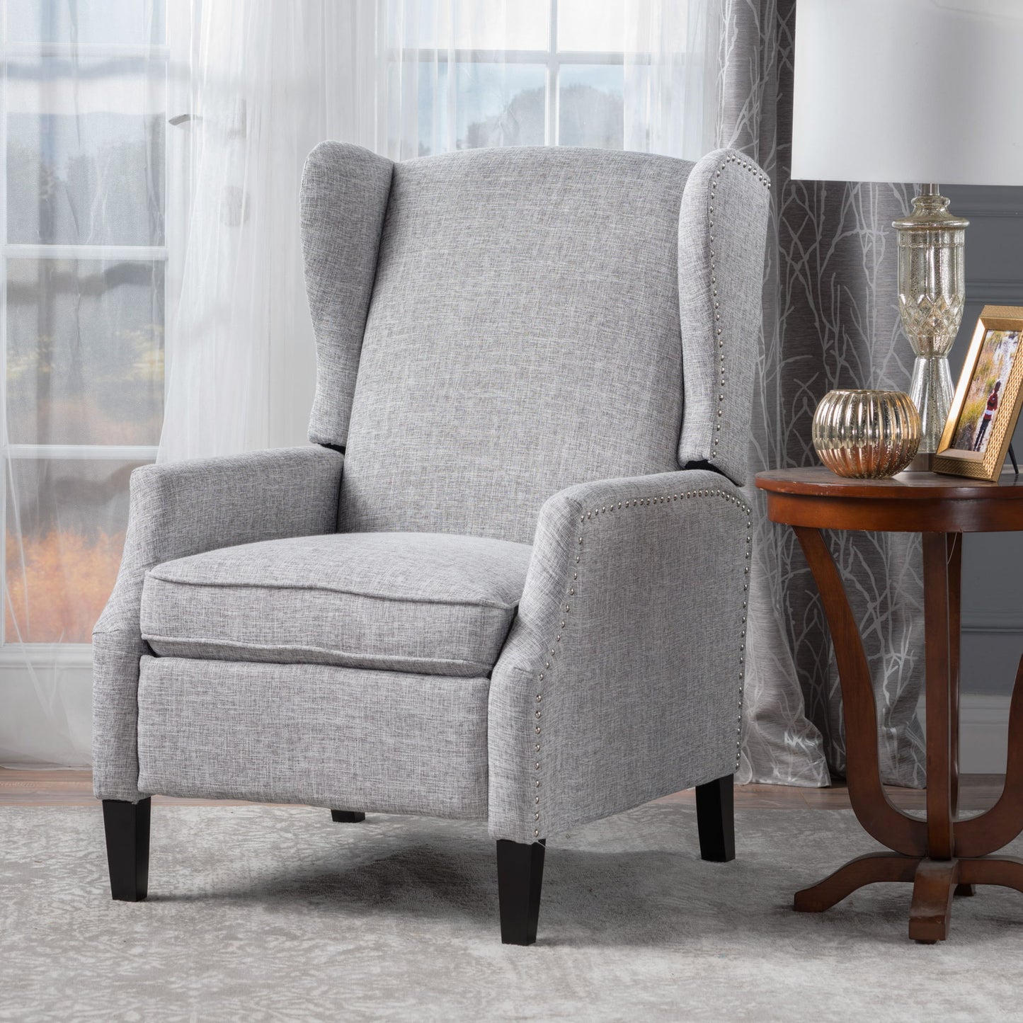 The Westeros Wing Back Fabric Upholstered Recliner in light gray sits next to a wooden side table with decor, set in a room featuring sheer curtains and large windows.