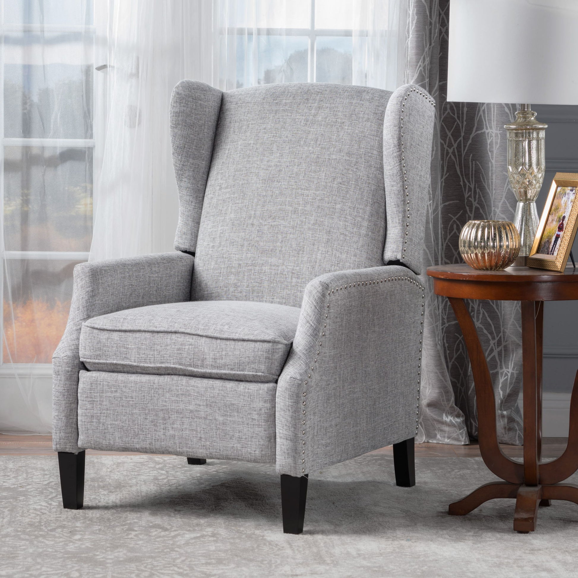 The Westeros Wing Back Fabric Upholstered Recliner in light gray sits next to a wooden side table with decor, set in a room featuring sheer curtains and large windows.