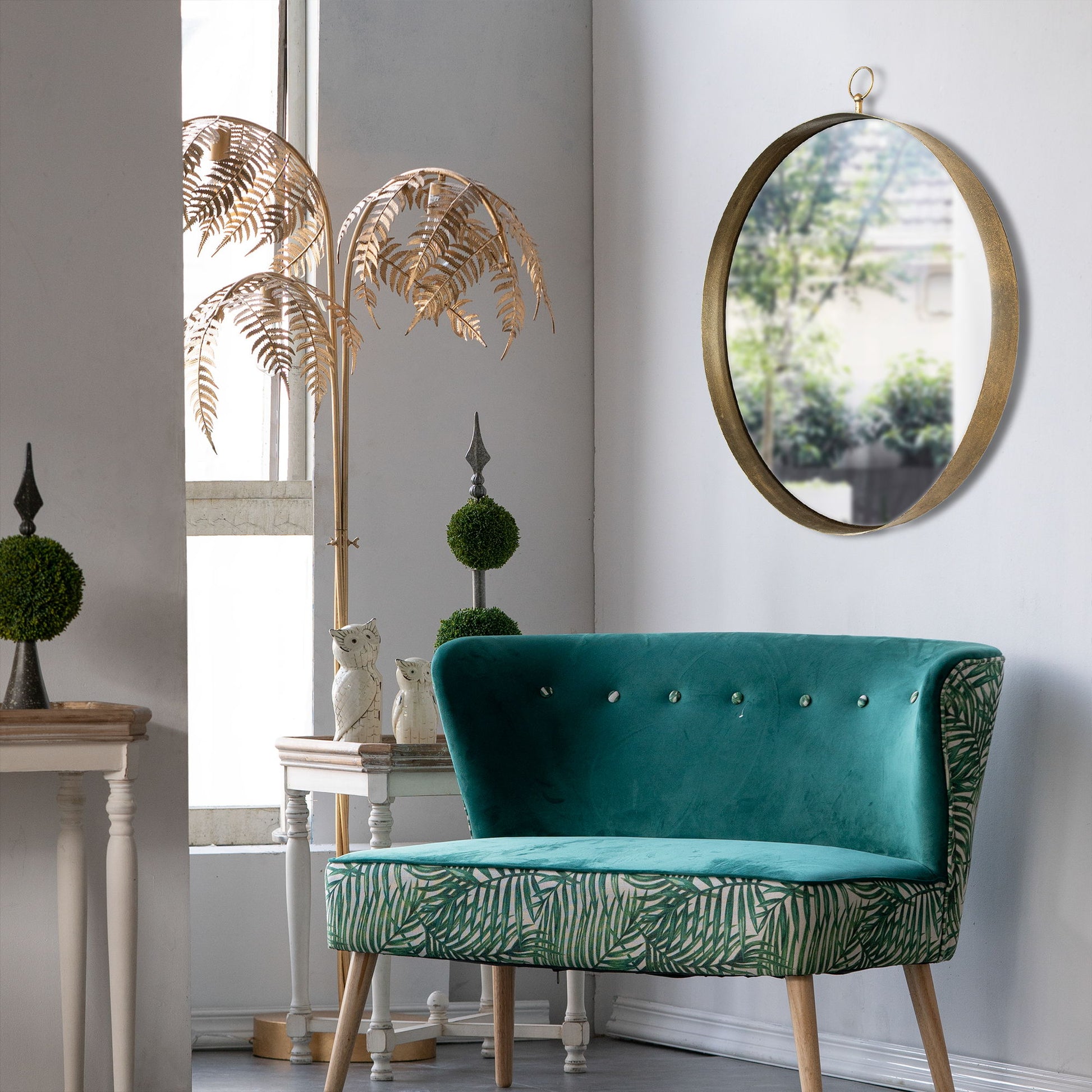 A stylish room features a turquoise velvet loveseat, potted plants, an owl figurine, a decorative gold palm tree by the window, and a Round Mirror with an iron frame hanging on the wall under soft natural light.