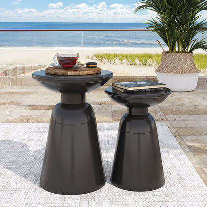 Two Serenita Outdoor Side Tables with a flared design sit on a beachside patio—one holds a tray with teacup and teapot, the other supports a tablet; a potted plant and ocean view provide the perfect backdrop.