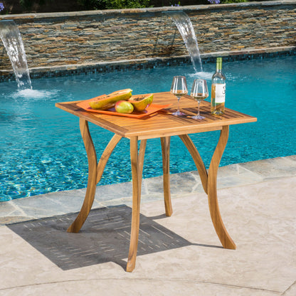 An Outdoor Rectangular Table With Curved Legs And Perpendicular Wooden Slats Table Top by a pool holds a tray with sliced papaya, white wine, and two glasses, set against a backdrop of a stone wall with water fountains.