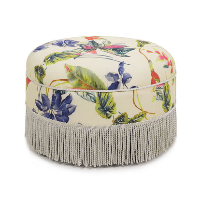 The Yolanda Round Upholstered Accent Ottoman features a cream background with vibrant floral and leaf patterns, finished with a long light gray fringe at the base.