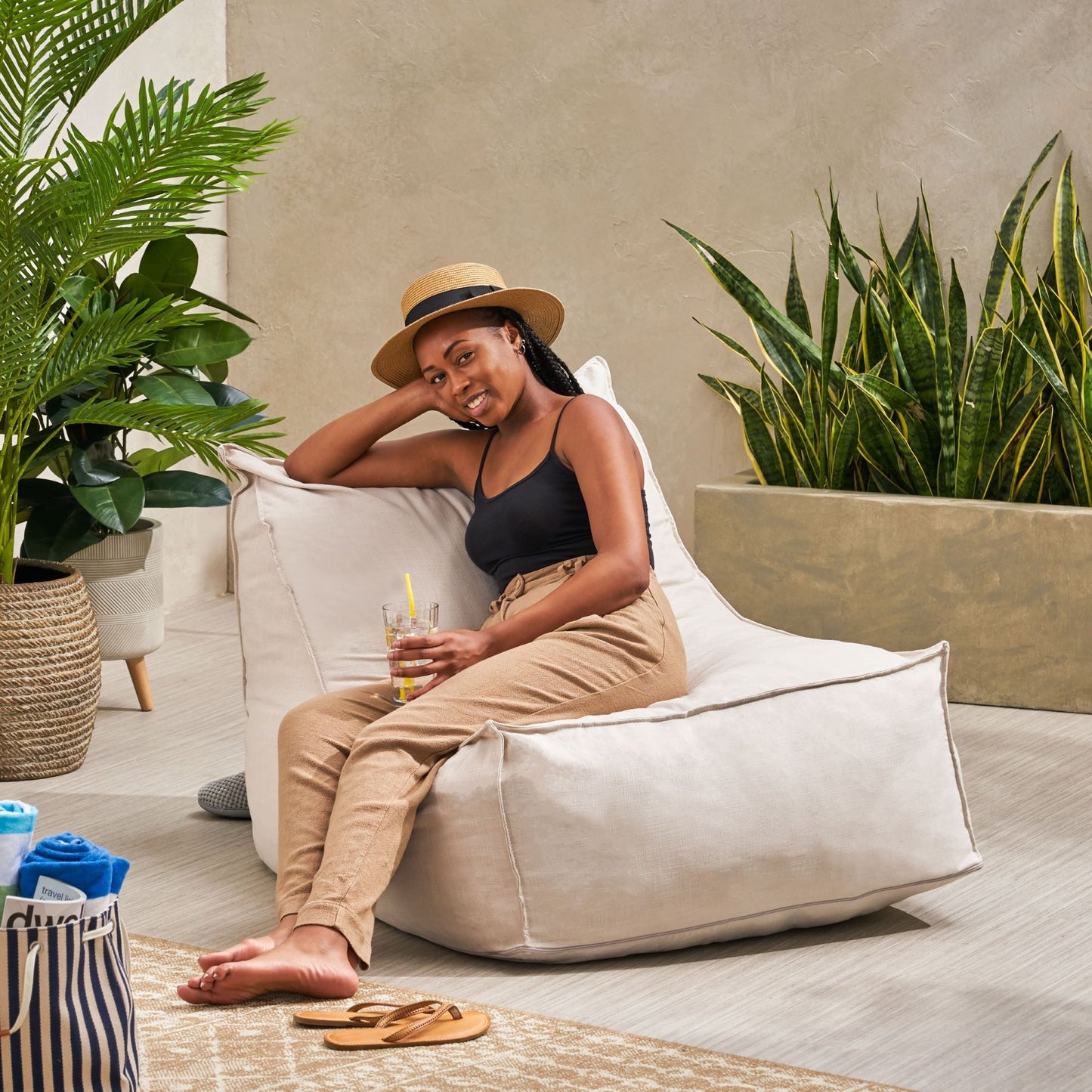 A woman in a hat, black tank top, and beige pants relaxes on the Ettie Water Resistant Fabric Bean Bag Chair. She holds a drink and smiles, surrounded by green plants in a bright, modern indoor space.